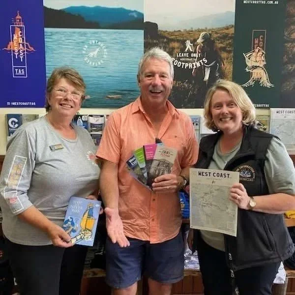 Three people smiling inside a store. The woman on the left has short hair and is holding brochures. The man in the middle has gray hair, wearing an orange shirt, holding flyers. The woman on the right has blonde hair, wearing a vest, and holding a map. Behind them are posters featuring outdoor scenes and travel posters for the West Coast.