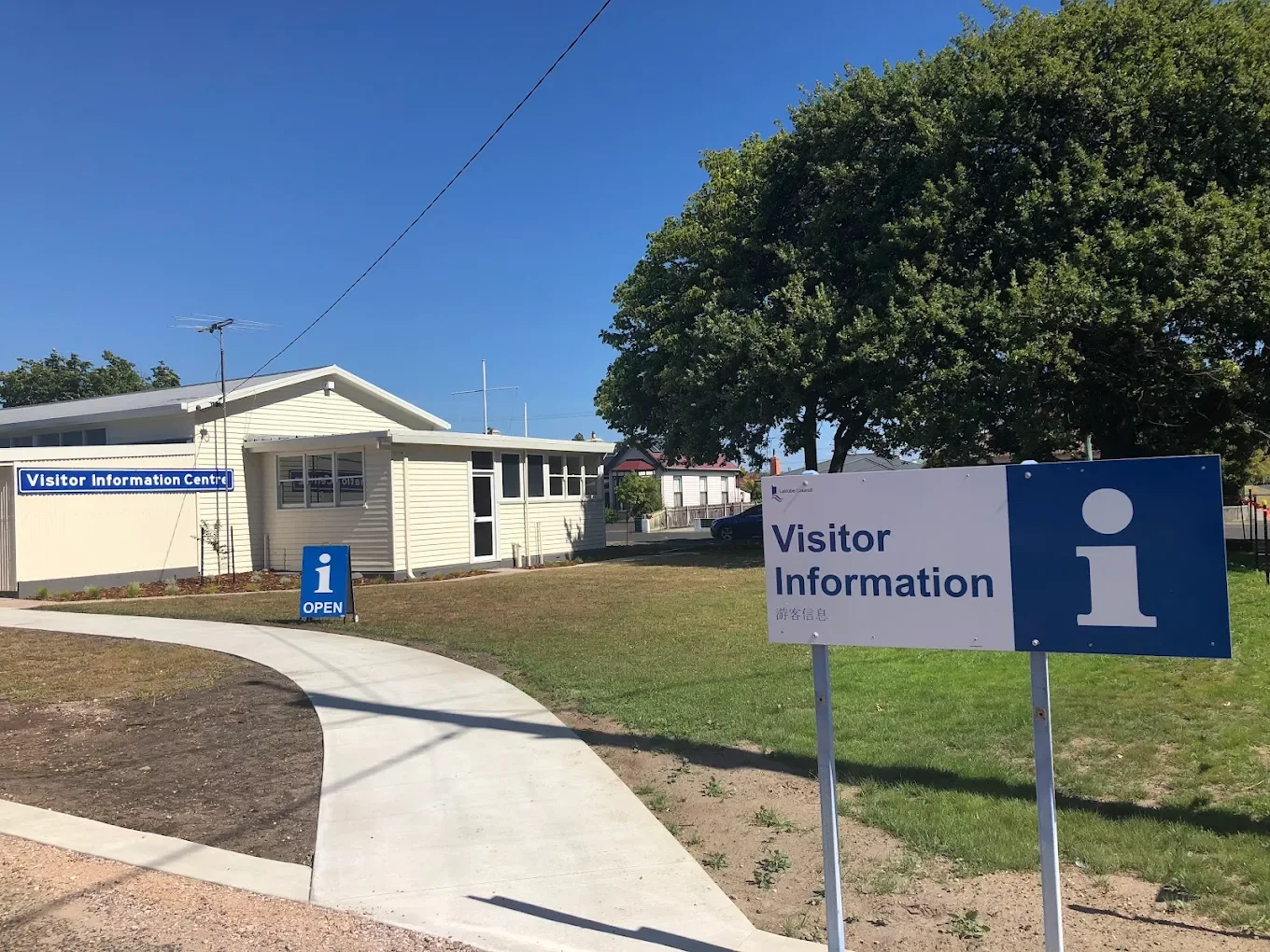 Visitor information center with signs, one large sign reading 'Visitor Information' with a white 'i' on a blue background, and another smaller sign with the same text and a blue 'i' on a white background, the building is white with windows, a sidewalk curves in front, and there are trees and a clear blue sky in the background.