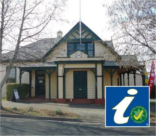 A historic two-story building with a gabled roof, a small porch, and a sign with a lowercase 'i' for information. The building has decorative trim and is surrounded by leafless trees and a sidewalk.