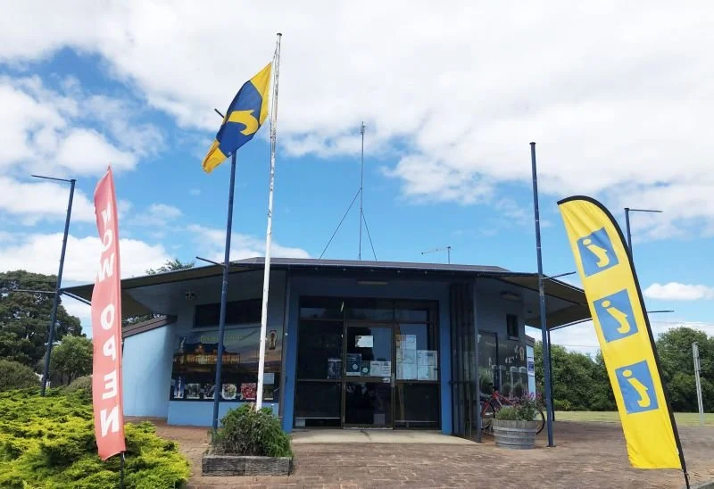 A small blue building with large glass windows and a flat roof, surrounded by three flags on tall poles, with trees and a cloudy sky in the background.