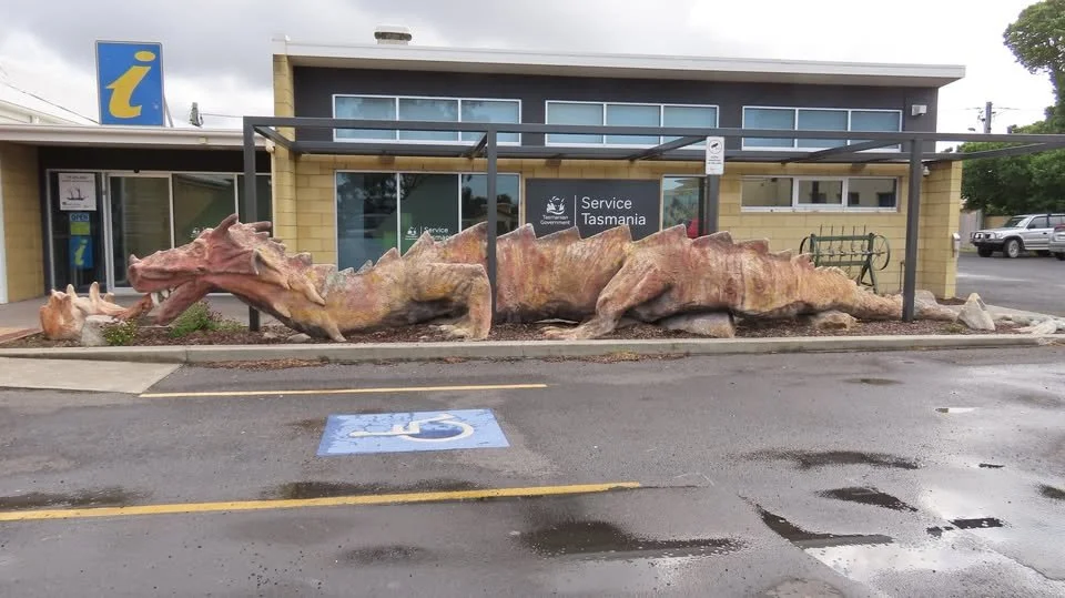 A large sculpture of a crocodile made of rocks placed in front of a building with glass windows. The building has a sign that reads 'Service Tasmania' and is located in a parking lot with spaces and a handicapped parking spot.