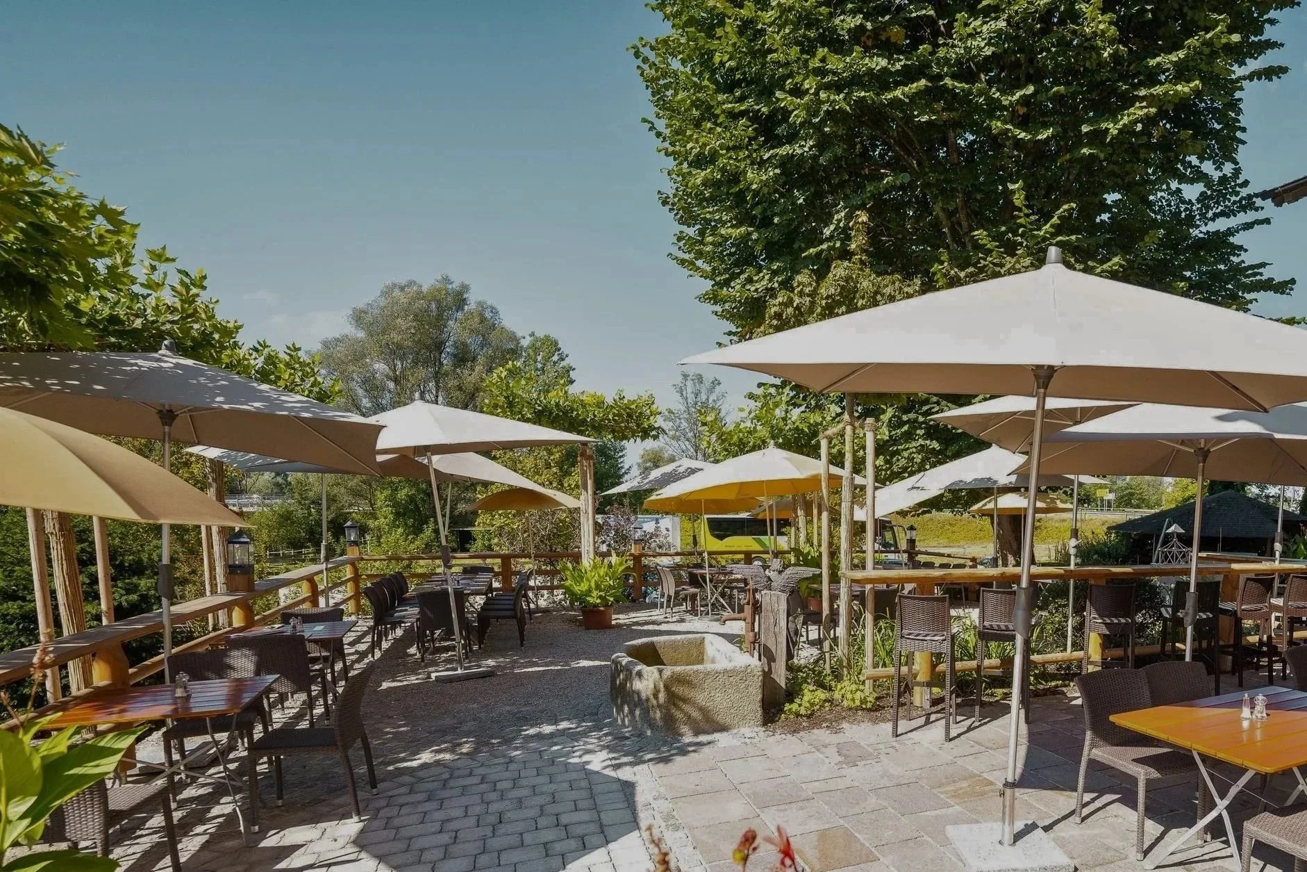 Ein Außenbereich eines Restaurants oder Terrassenbereich mit mehreren Tischen und Stühlen, Sonnenschirmen und viel Grün durch Bäume, bei sonnigem Wetter