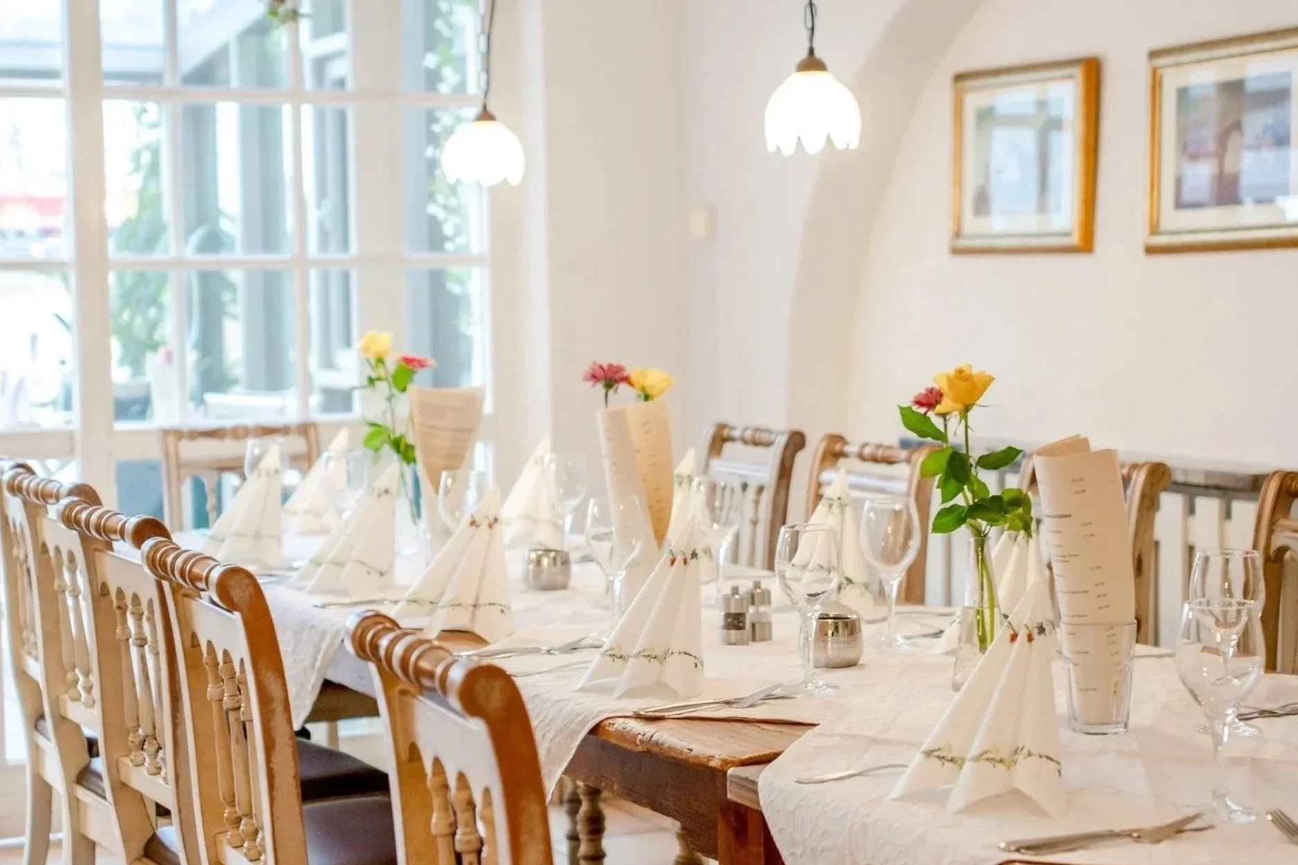 Ein festlich gedeckter Esstisch mit weißen Servietten, Menükarten, Gläsern und kleinen Kerzen. Die Tafel ist mit kleinen Vasen mit bunten Blumen dekoriert. Im Hintergrund sind große Fenster und einige Bilder an der Wand.