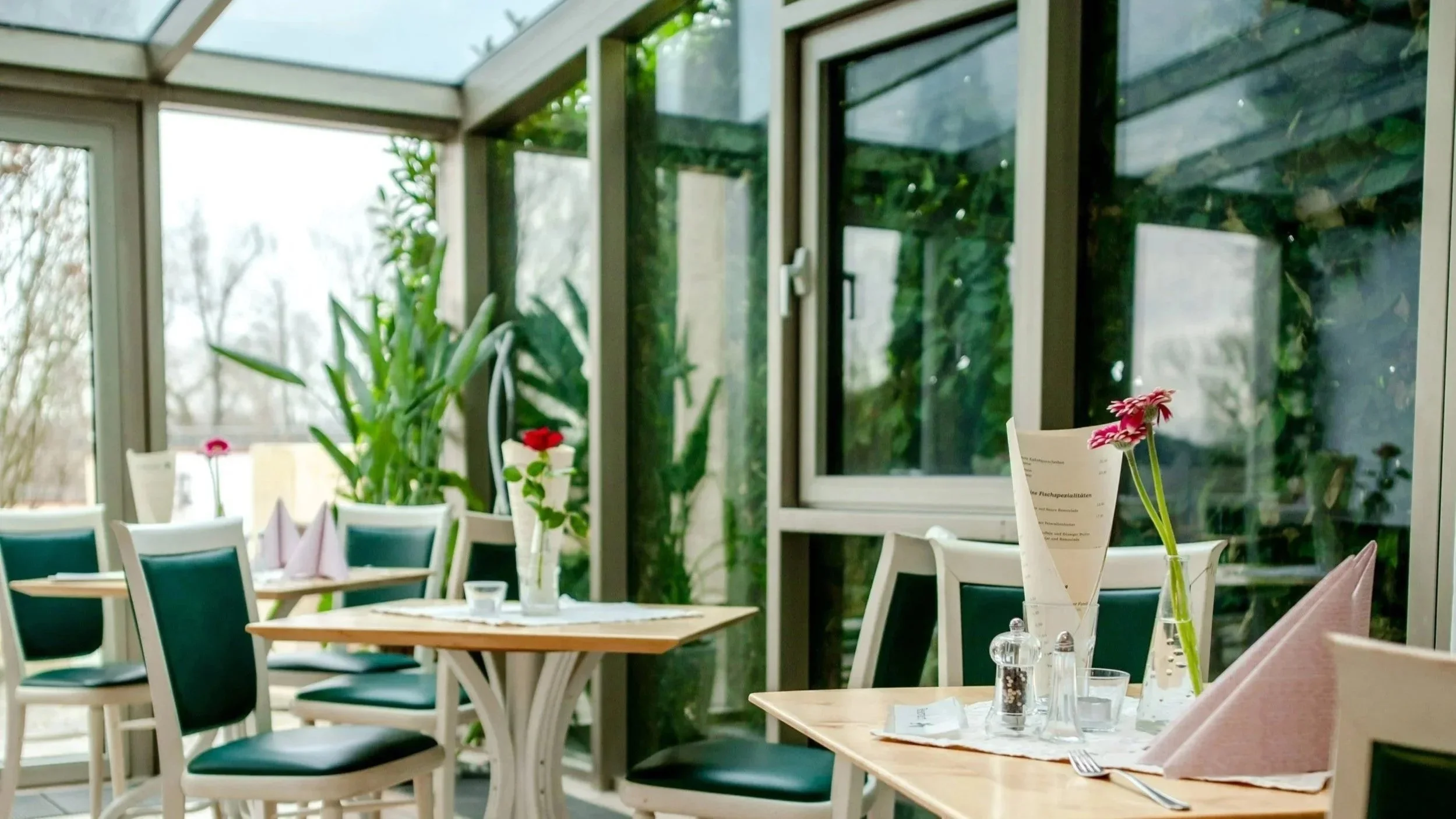 Innenansicht eines sonnigen Restaurants mit Tischen, Stühlen, Blumen in Vasen und großen Fenstern mit Blick ins Grüne.