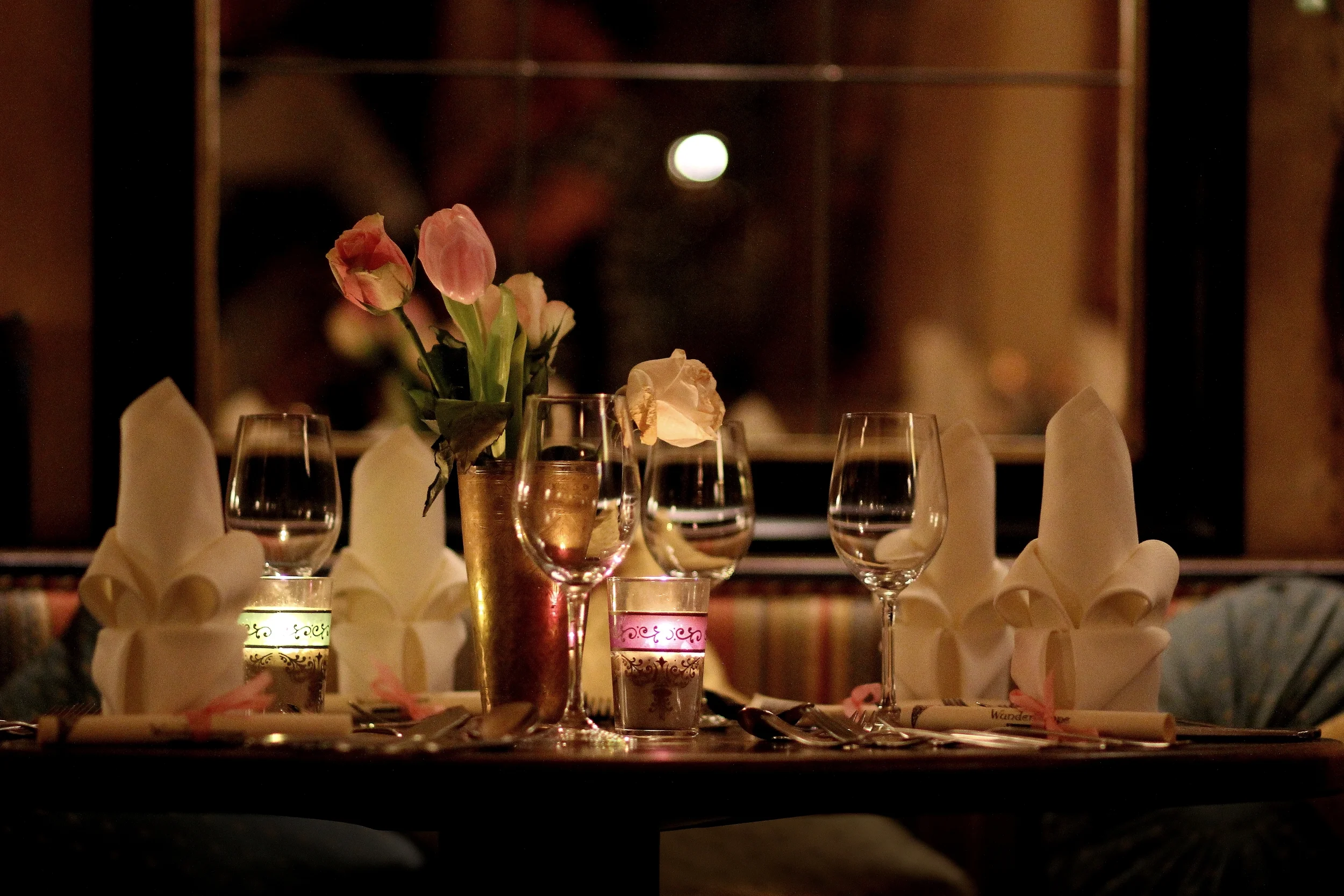 Ein gedeckter Tisch mit Blumen in einer Vasen, Weingläsern, Kerzen und mit Papptellern in einem Restaurant. Es ist abends, mit warmem Licht und einer dunklen Fensteransicht.