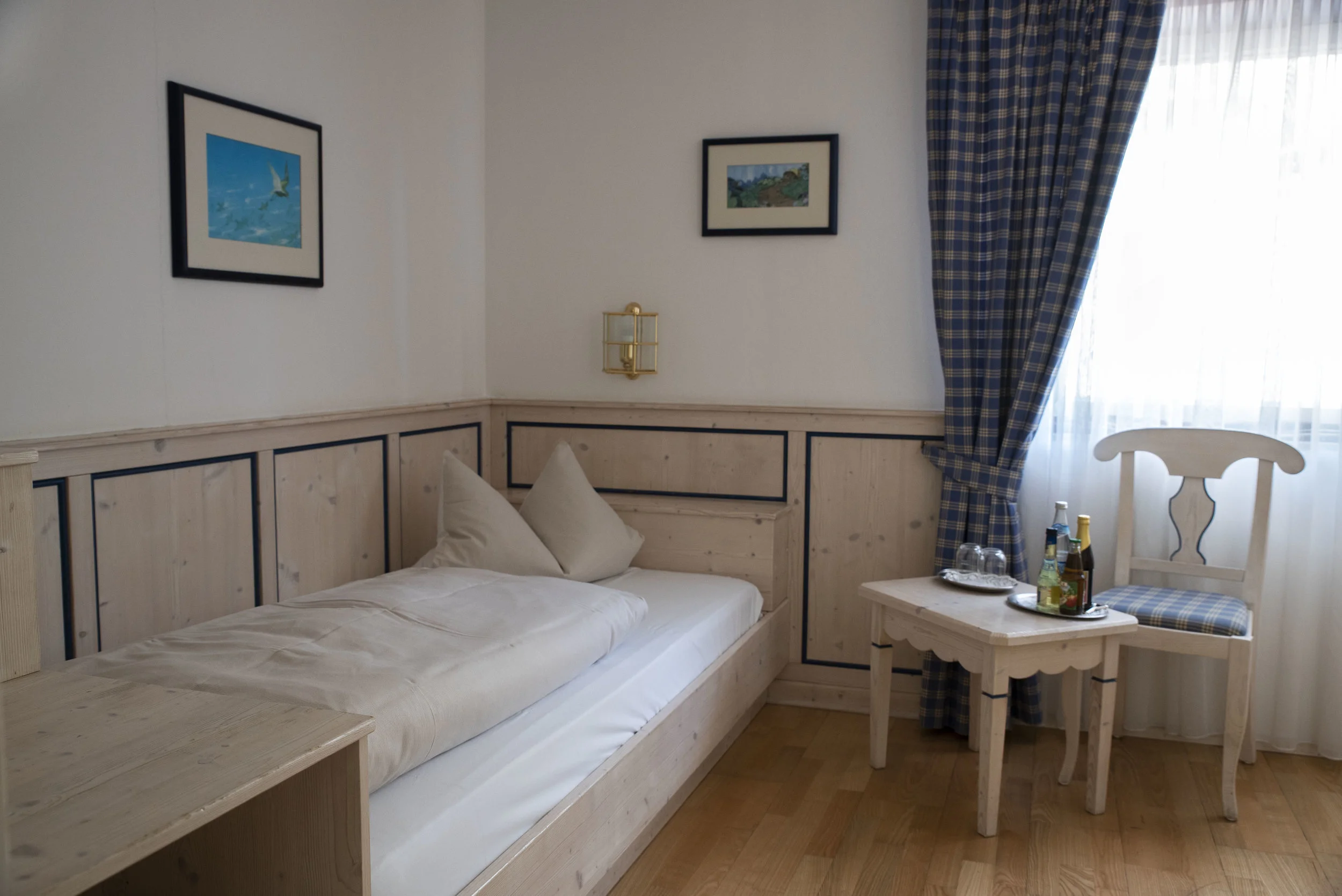 Ein Hotelzimmer mit weißem Bett, zwei weißen Kissen, einer kleinen Holzkiste neben dem Bett, einem runden Tisch mit Getränken, einer Holzstuhle mit blau-karierter Sitzfläche, beigen Wänden mit Bilderrahmen, Vorhängen mit blauen Karos und einem großen Fenster.