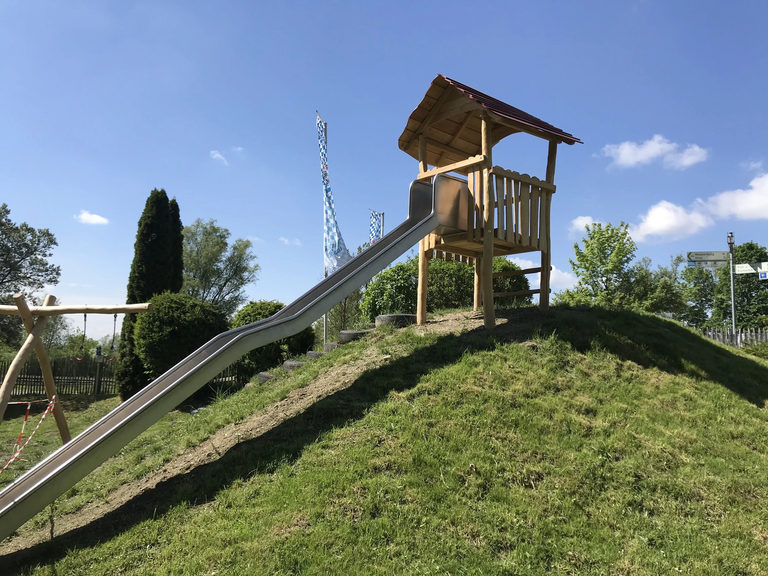 Ein Spielplatz mit einer Rutsche, einem Baumhaus, einem Holzschaukel und grüner Vegetation unter blauem Himmel.