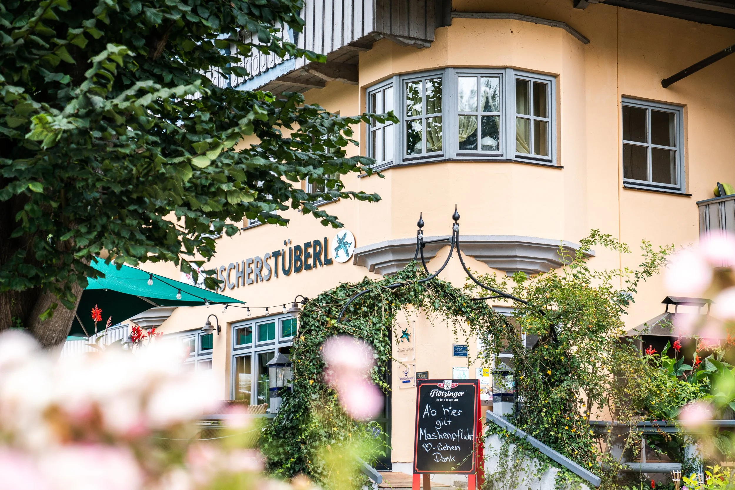 Mehrstöckiges Gebäude mit gelben Wänden, großen Fenster und Balkonen, umgeben von grünen Bäumen und Blumen, mit einer Tafel vor dem Eingang, die Werbung und Hinweise enthält.