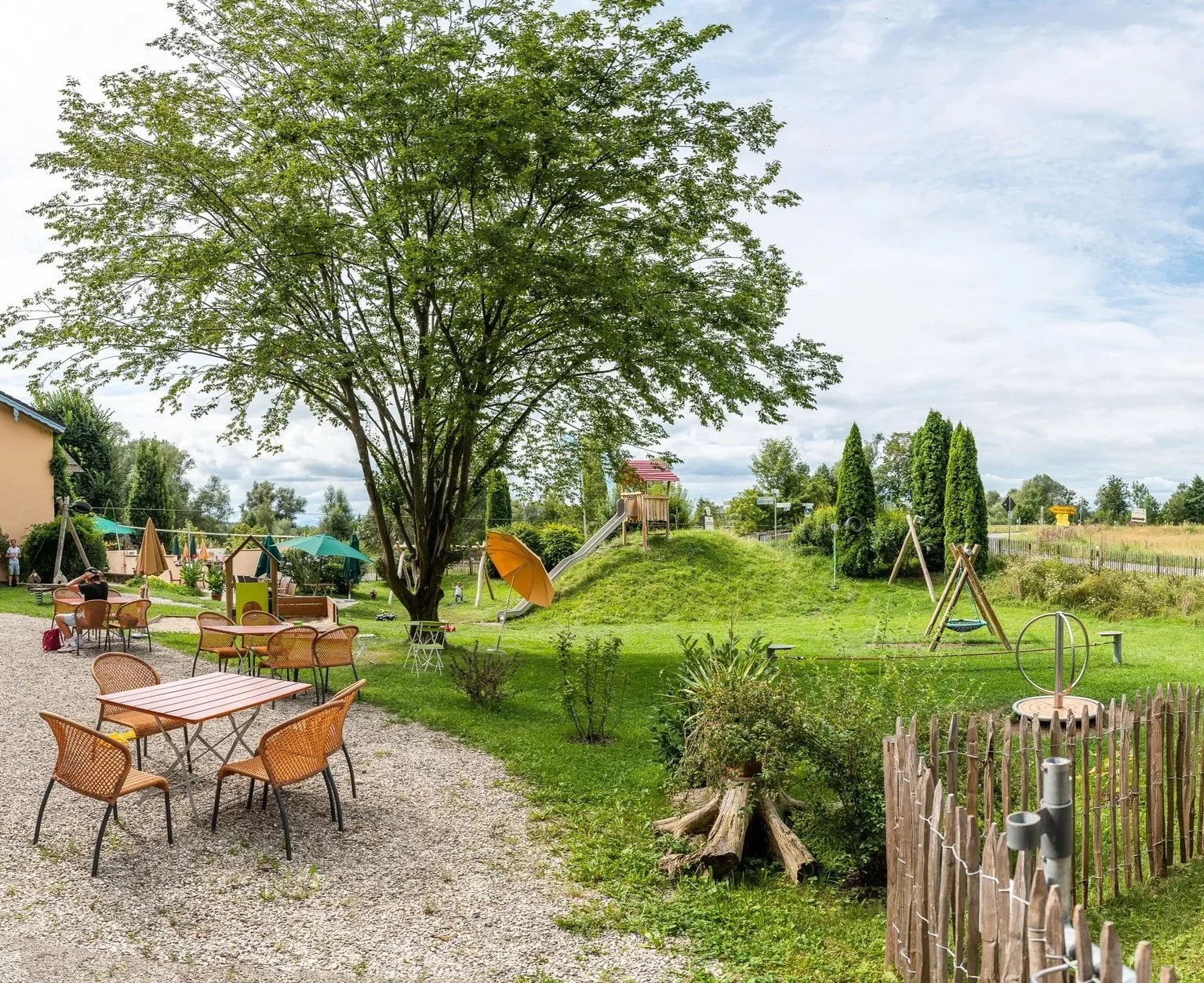 Ein Kinderspielplatz im Freien mit Rutsche, Schaukel und Bäumen, umgeben von einem Garten mit Tischen und Stühlen, in einer ländlichen Umgebung bei bewölktem Himmel.