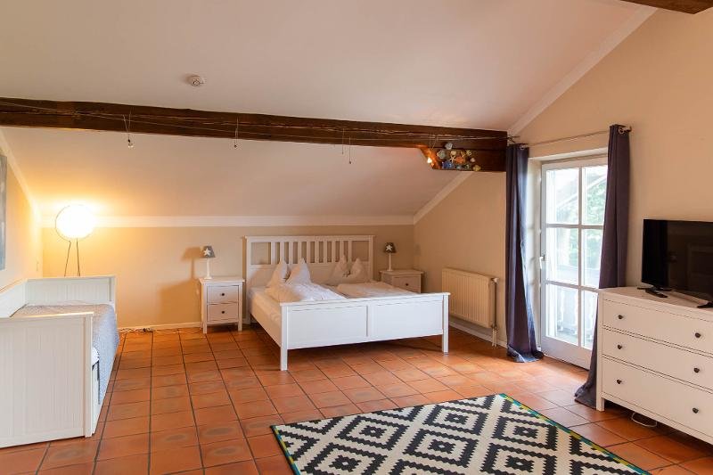 Schlafzimmer mit Doppelbett, Nachttischen, Kommode, Sessel, Teppich, und Zugang zu einem Balkon mit Tür. Warme Beleuchtung, Holzbalken an der Decke, und Terrakottaboden.