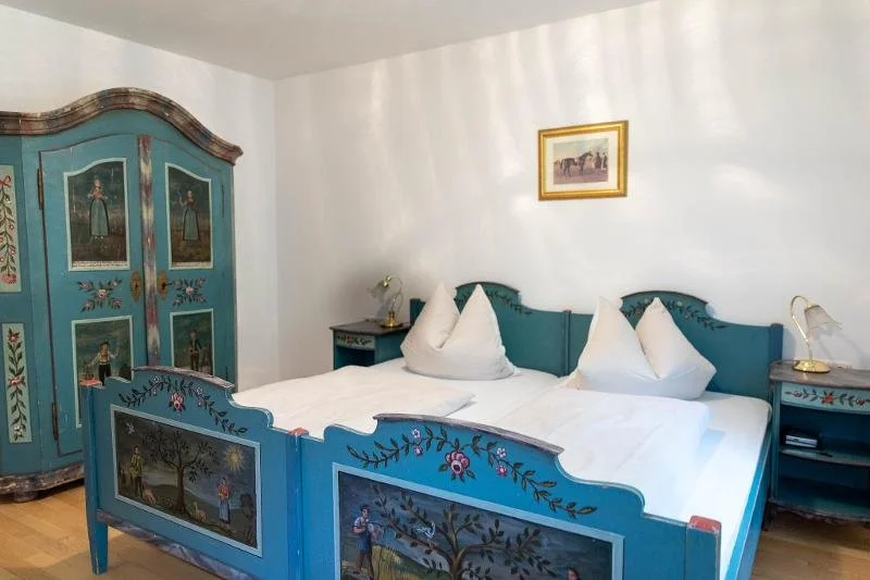 Ein Schlafzimmer mit blau gestrichenem, handbemaltem Holzbettrahmen und Schränken, weißen Bettdecken und Kissen, Wandbildern, Holzboden.