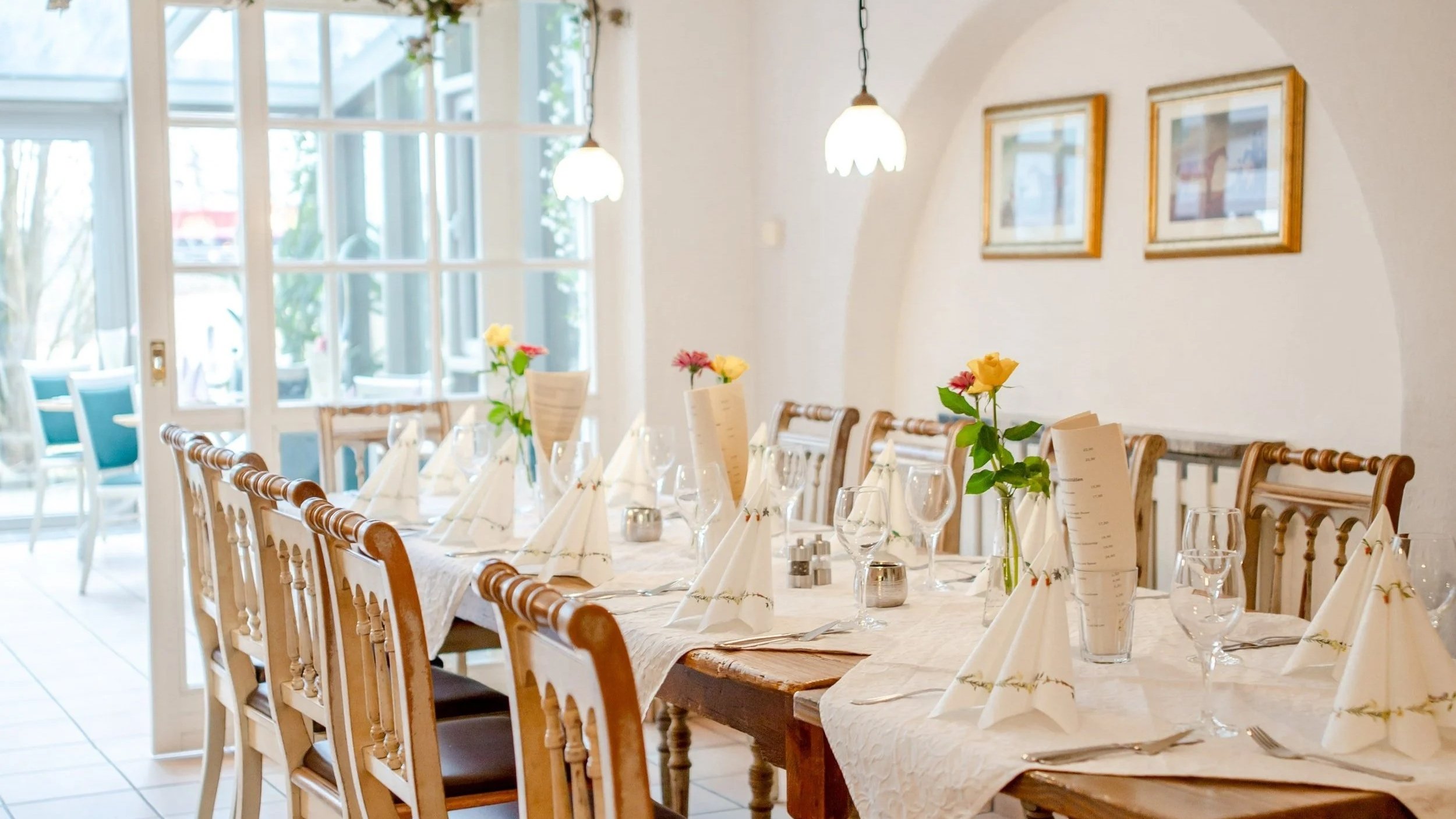 Ein festlich gedeckter Tisch in einem hellen Restaurant mit Blumen in Vasen, Weingläsern, Besteck und Menükarten, mit großen Fenstern im Hintergrund.