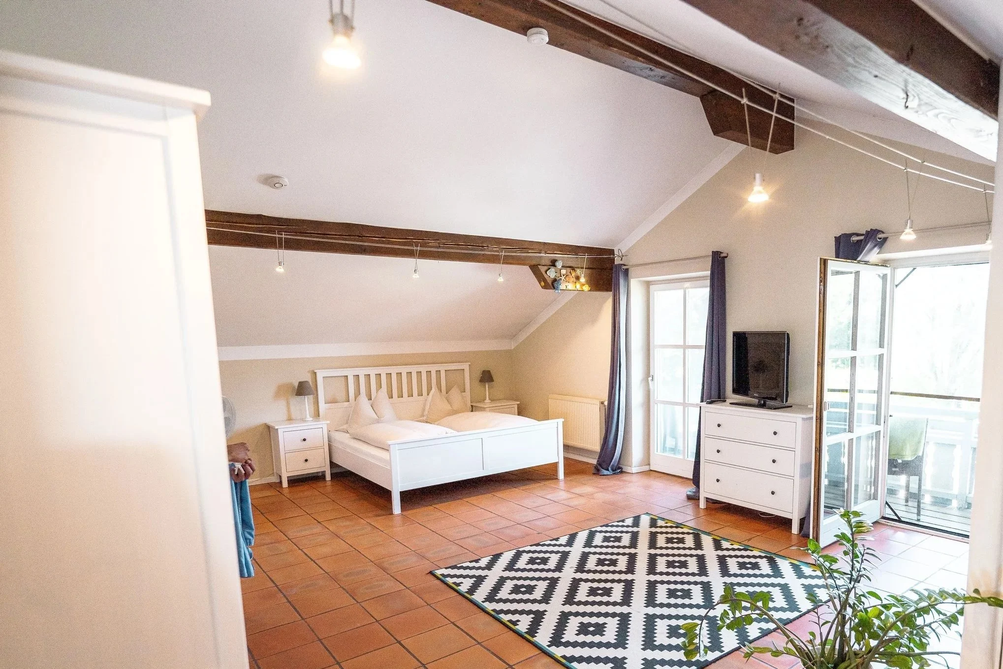 Ein Schlafzimmer mit weißen Möbeln, einem Bett, Nachttischen, einem Fernseher auf einer Kommode, einem offenen Balkon mit Türen und Fenster, dunklen Vorhängen, einem schwarzen und weißen Teppich, Holzbalken an der Decke und terrakottafarbenen Fliesen auf dem Boden.