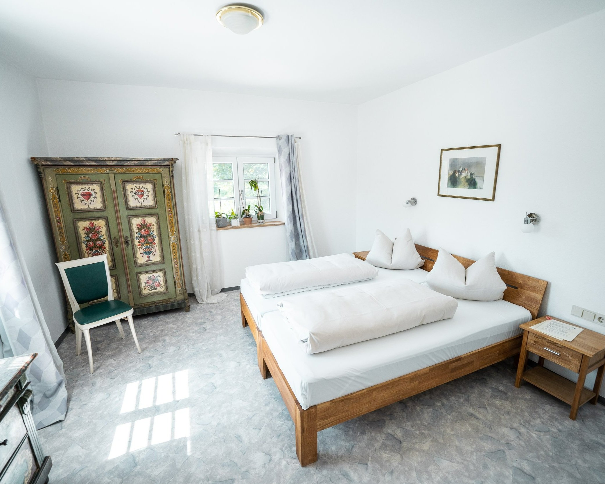 Schlafzimmer mit Holzbett, weißen Kissen und Decke, alten grünen Türe mit Blumenmalerei, Fenster mit weißen Vorhängen und einigen Pflanzen, Wandbild, Wandlampen, grauer Boden