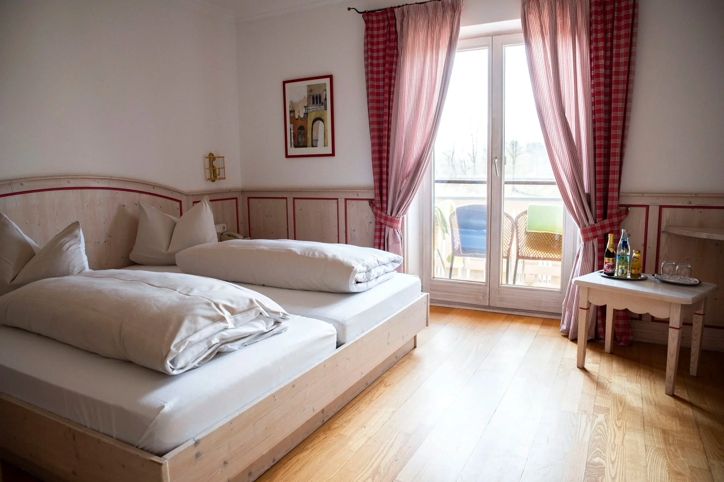 Ein Hotelzimmer mit zwei Betten, großen Fenstern mit roten karierten Vorhängen, einem kleinen Tisch mit Getränken, einer Wanddekoration und Blick auf den Balkon mit Stühlen.