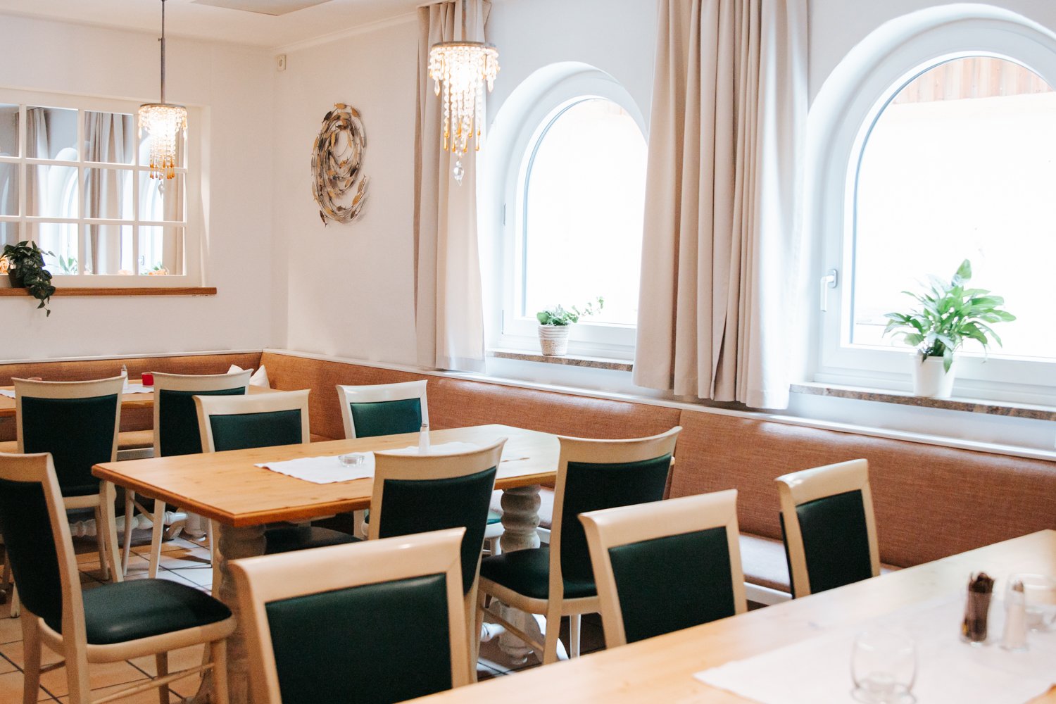 Gemütliches Restaurant mit Holztischen, grünen und weißen Stühlen, zwei großen Fenstern mit Vorhängen, Pflanzen auf den Fenstersimsen, Kandelabern an der Decke und Wanddekorationen.