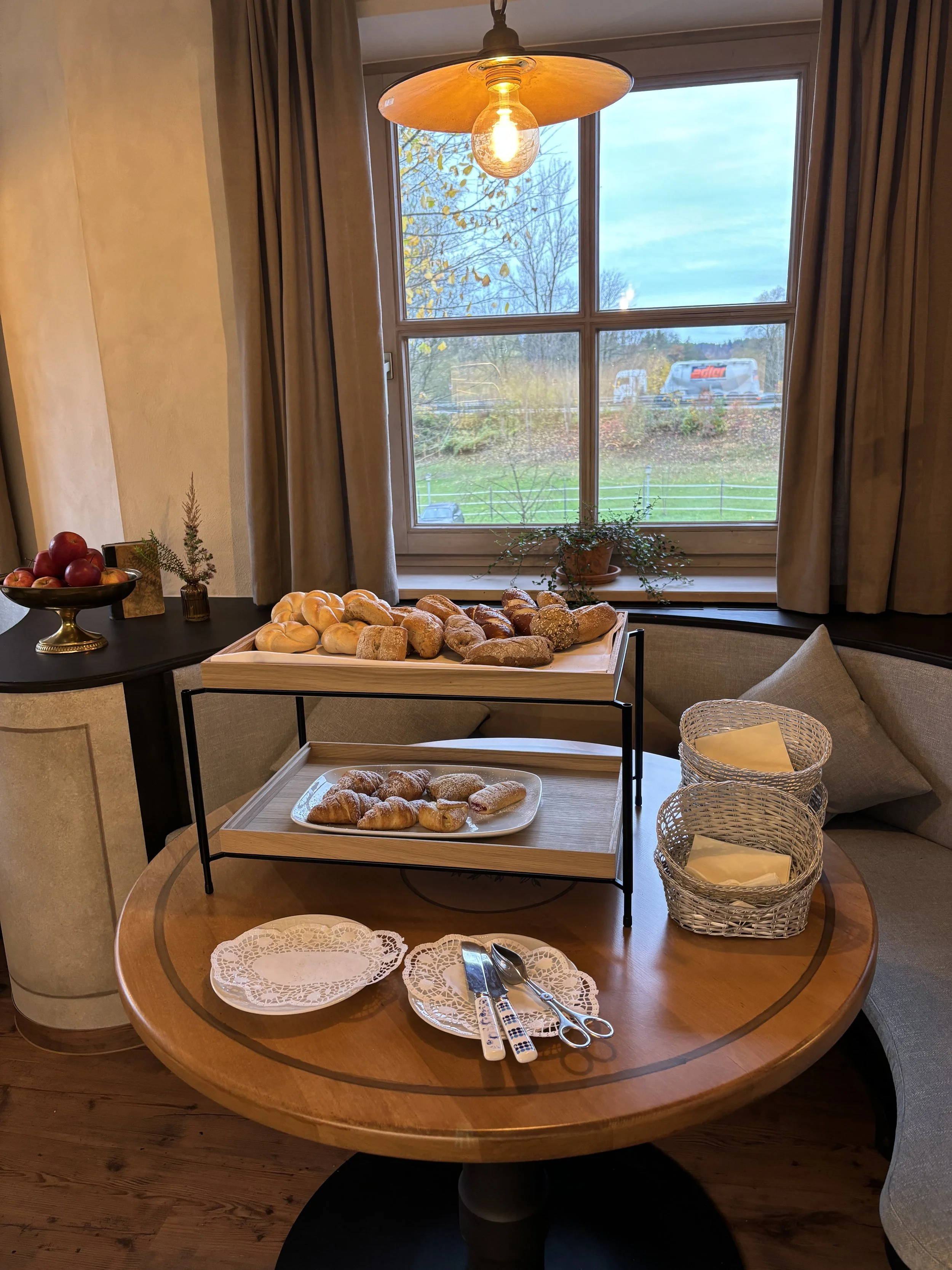 Ein runder Holztisch mit einer Tablett voller Brötchen, zwei weiße Teller und Besteck, zwei dekorative Körbe mit Servietten, auf der Fensterbank eine Topfpflanze, im Hintergrund ein Fenster mit Vorhängen und eine Aussicht auf Bäume und Straßen.