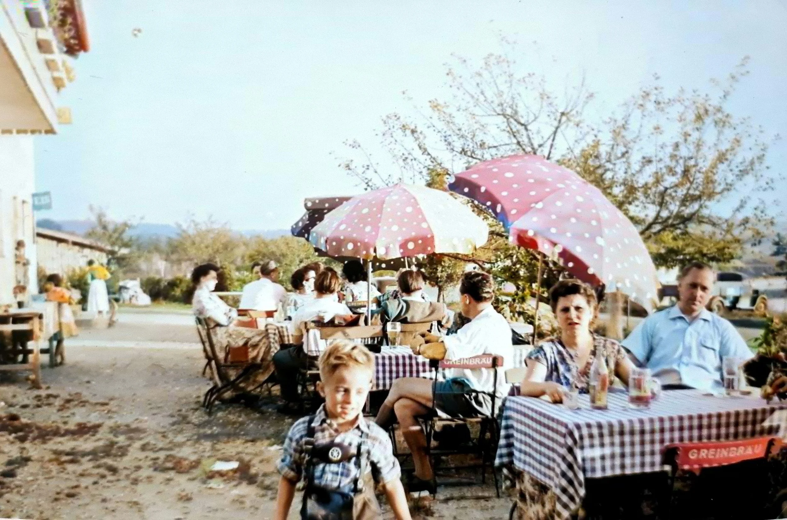 Menschen sitzen bei einem Treffen im Freien unter rosa und weißen Sonnenschirmen. Es gibt Bäume mit herbstlichen Blättern und eine klare Himmel im Hintergrund.