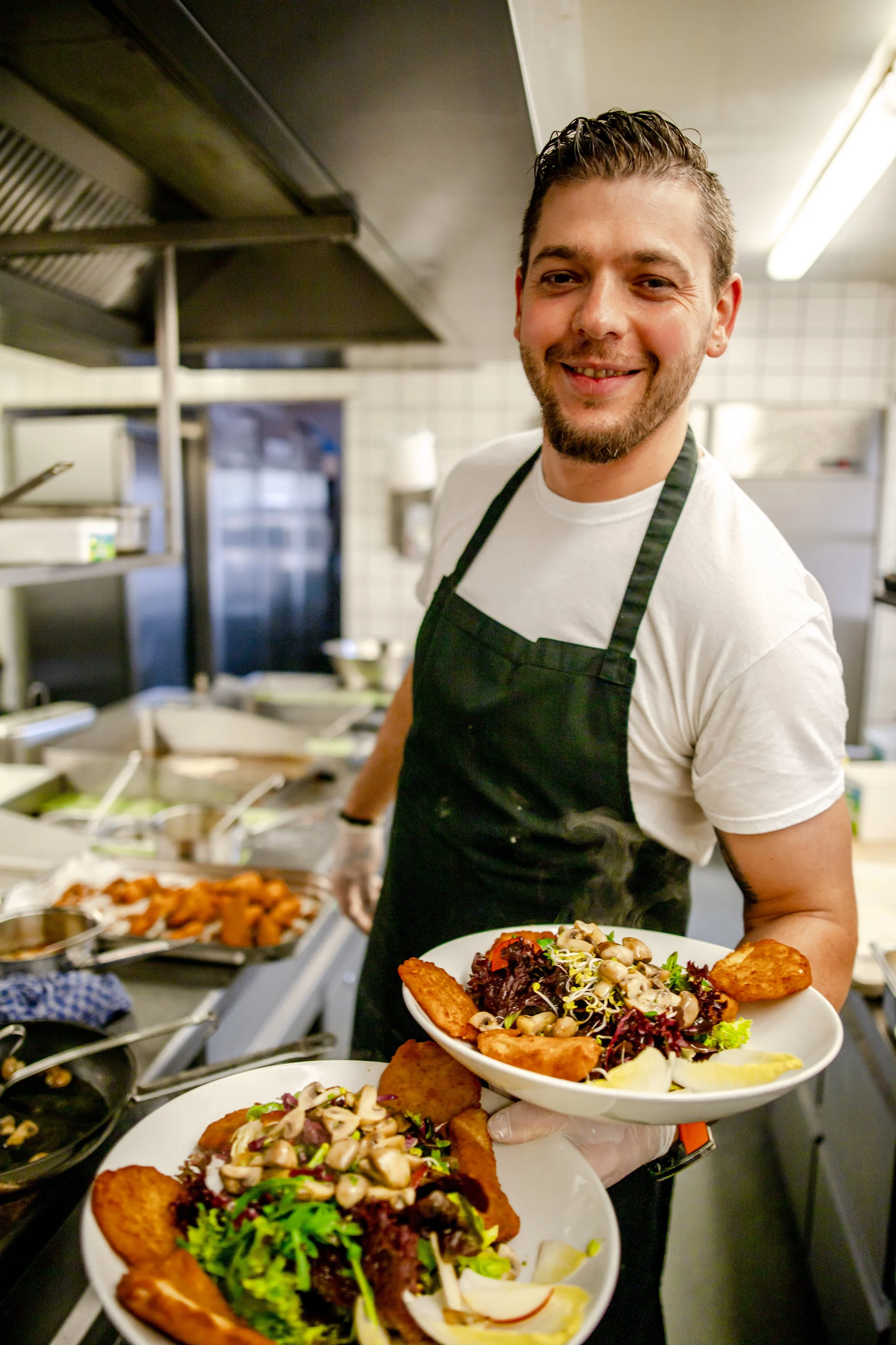 Ein Mann im schwarzen Kochschurz und weißen T-Shirt, der in einer Küche Essen serviert, hält einen Teller mit Salat und frittiertem Fisch. Im Hintergrund sind weitere Gerichte und Küchengeräte sichtbar.
