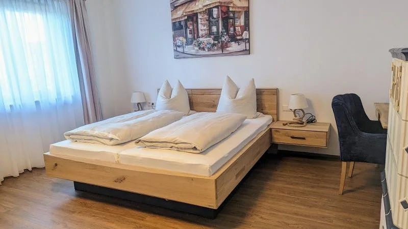 Schlafzimmer mit Doppelbett, Nachttischen, Nachttischlampen, einem Wandbild, einem Stuhl und einem Fenster mit Vorhängen.