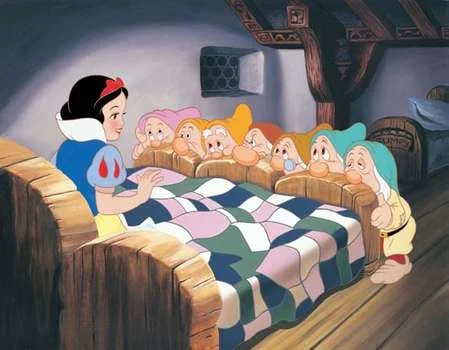Snow White sits in bed, surrounded by the seven dwarfs in a cozy wooden cottage.
