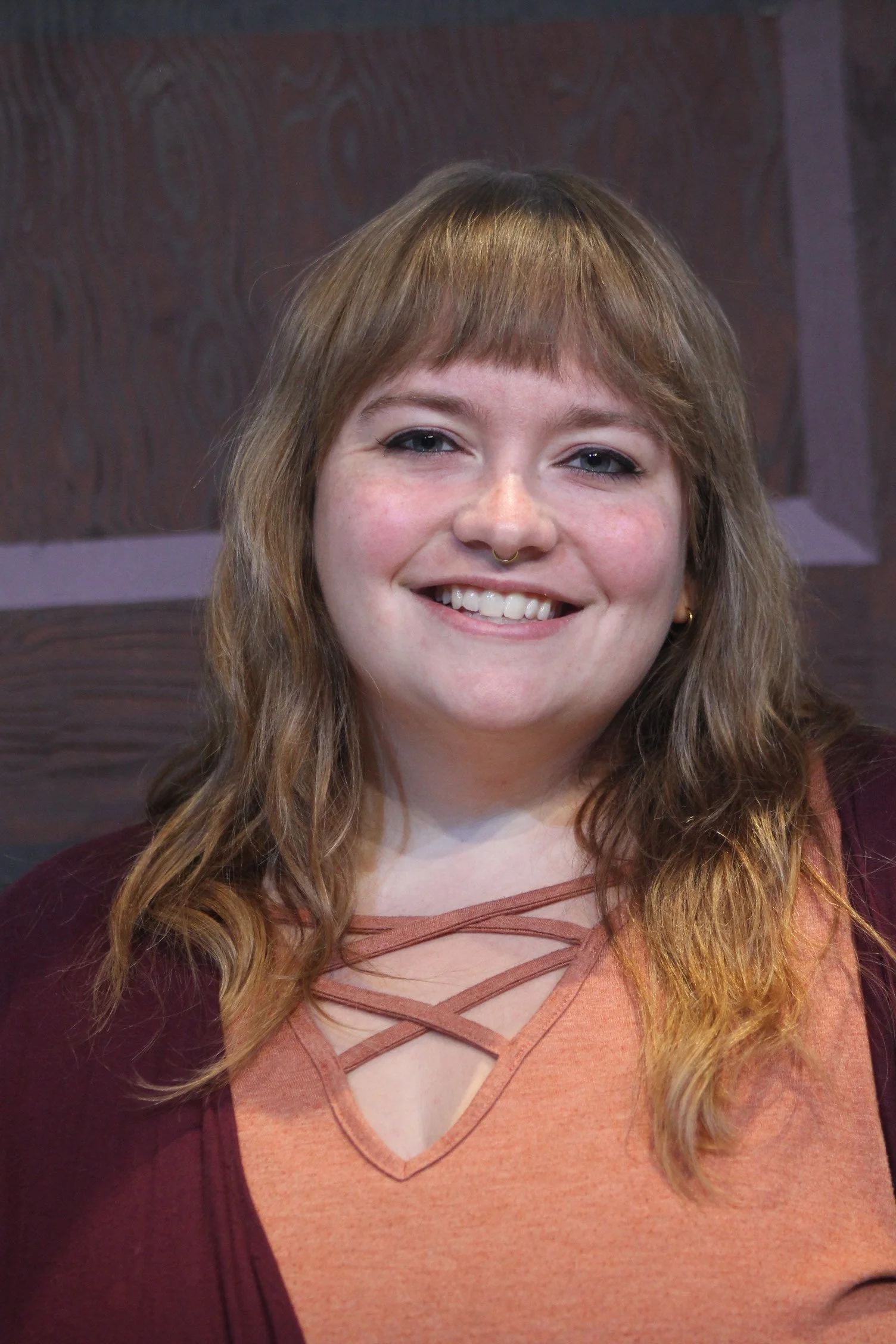 Image of Caucasian woman with light brown wavy hair and bangs wearing an orange blouse with criss-cross detail