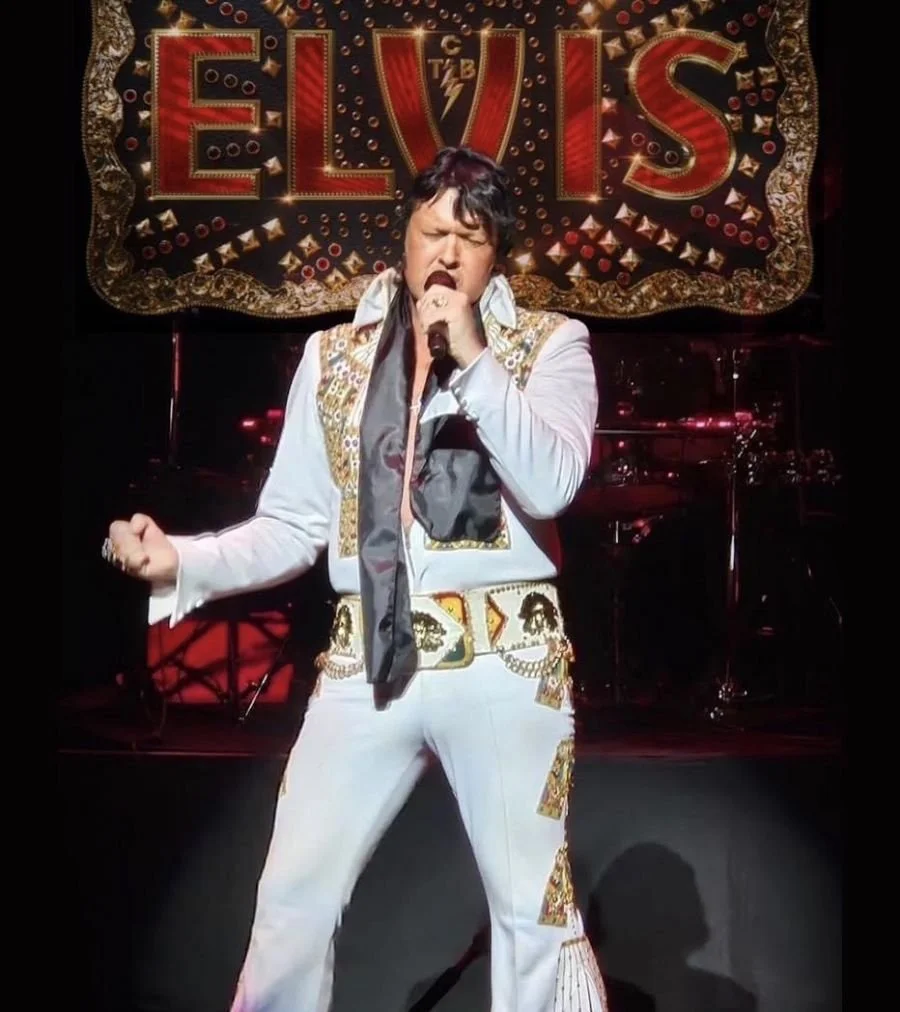 Singer dressed as Elvis in a white costume holding a microphone with the word "Elvis" emblazoned behind him.