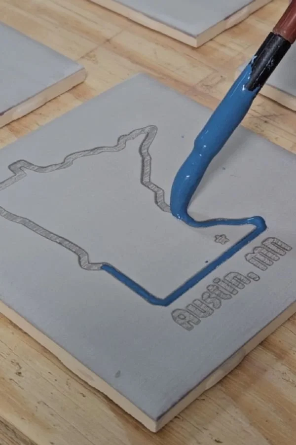 Image of clay tile being glazed with blue glaze in an outline of the shape of the state of Minnesota
