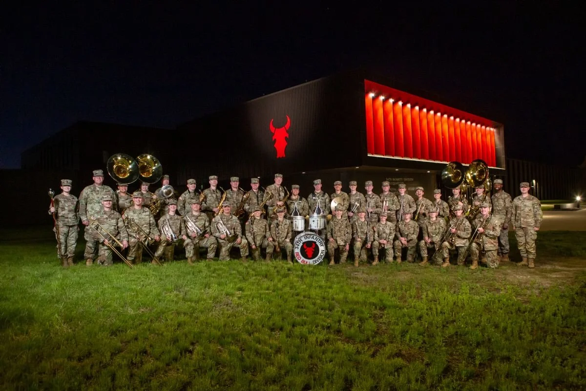 Image of 34th Infantry Division Army National Guards with their instruments in two rows in front of a red lit building illuminated with a red bull icon