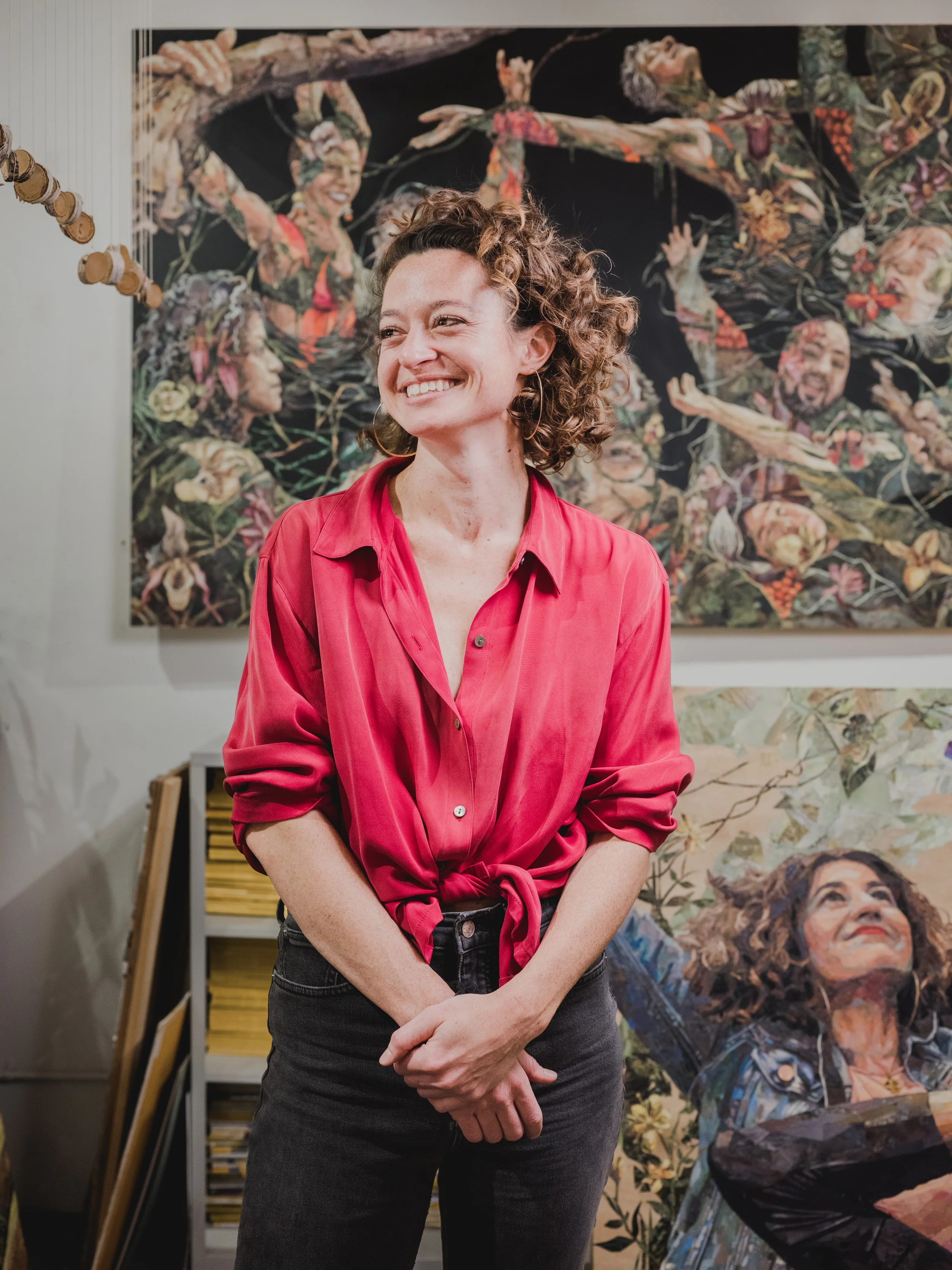 Image of female presenting person standing in front of artwork with brown curly hair and a red button down blouse smiling