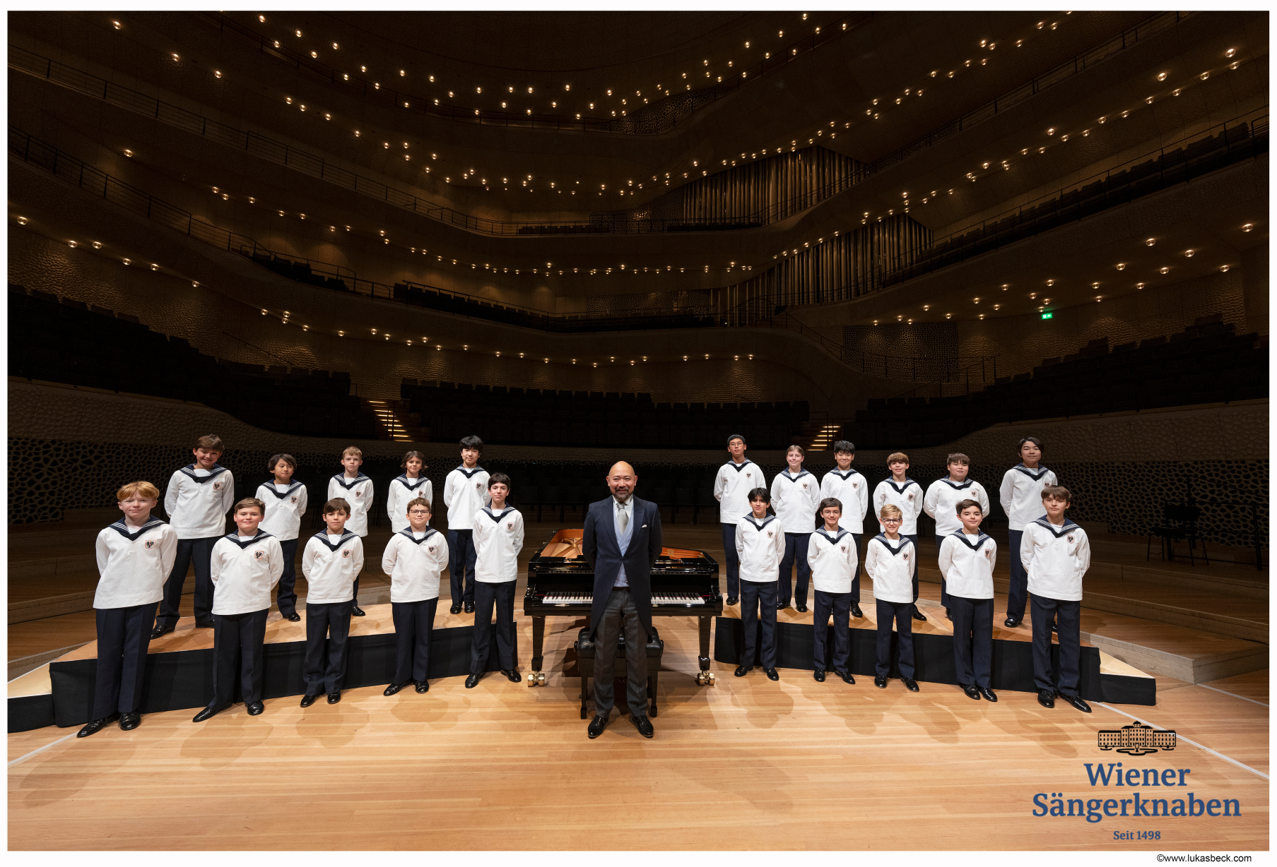 Image of choir on a stage with their conductor standing in front of a piano in the middle of the two rows,  in a large concert hall.