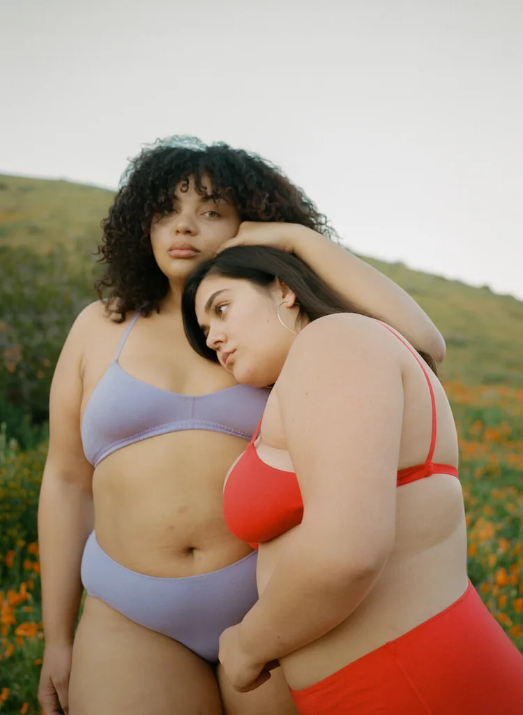 Two models in red and lilac underwear lean together, one resting a hand on the other’s hair.