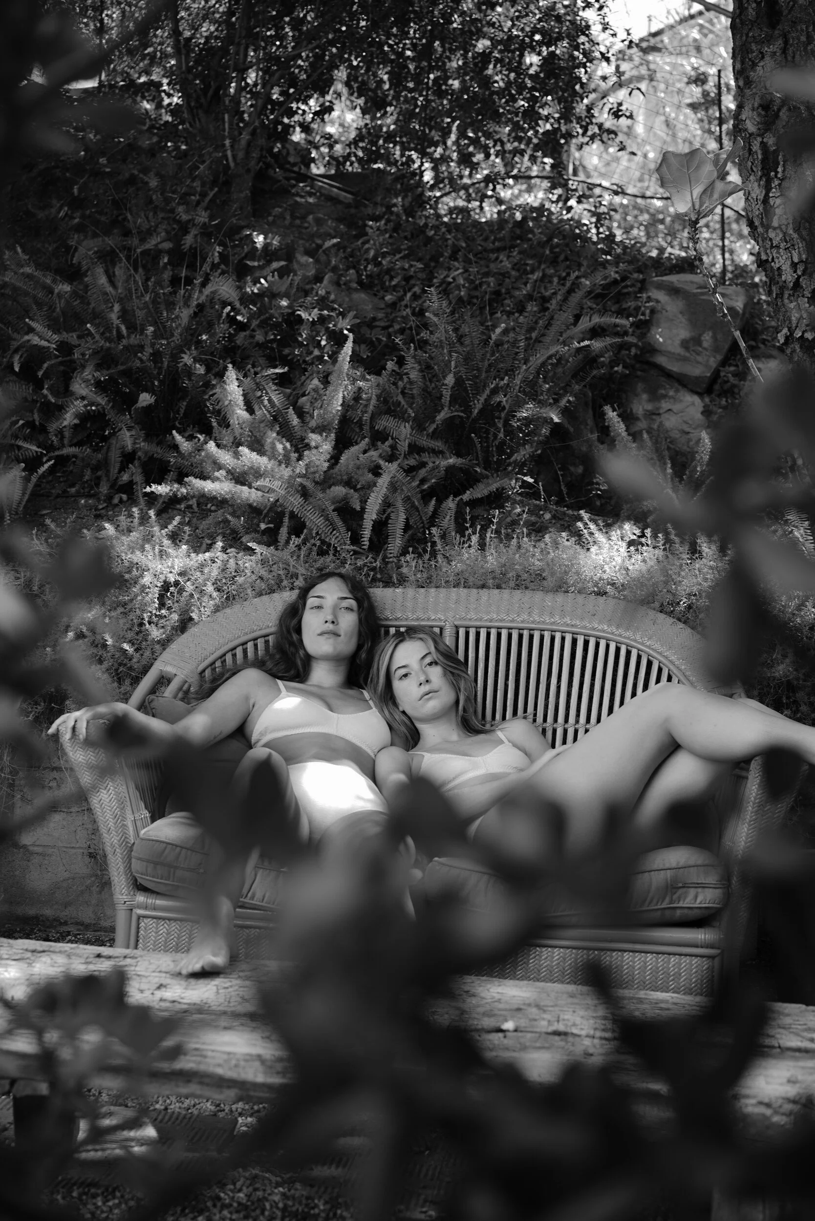 In black and white, two models in underwear recline together on a curved garden sofa surrounded by ferns.