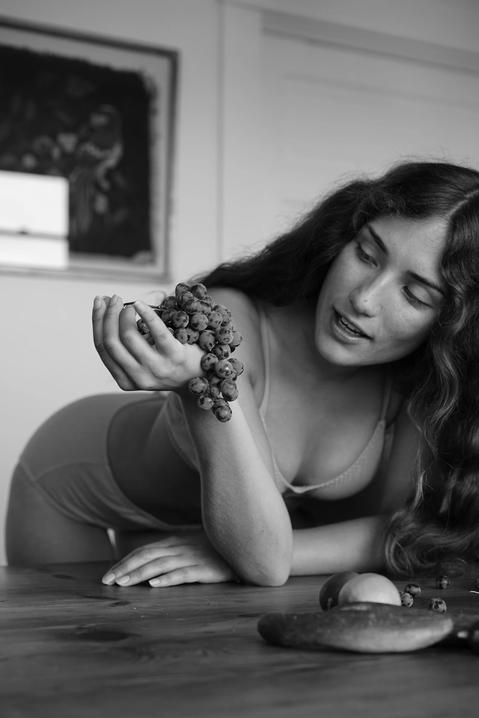 In black and white, a model in underwear leans over a wooden table holding a bunch of grapes.