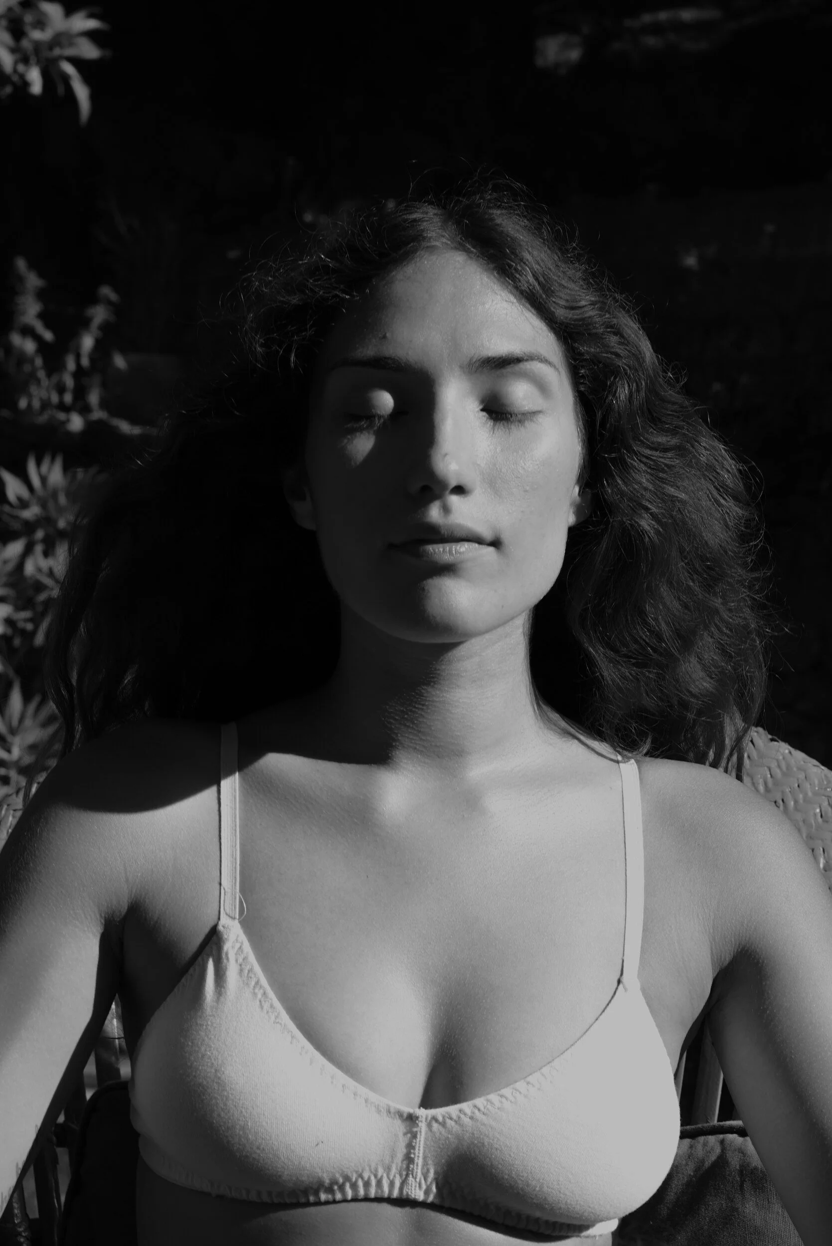 In black and white, a model in a light bra sits outdoors with eyes closed and sunlight across their face.