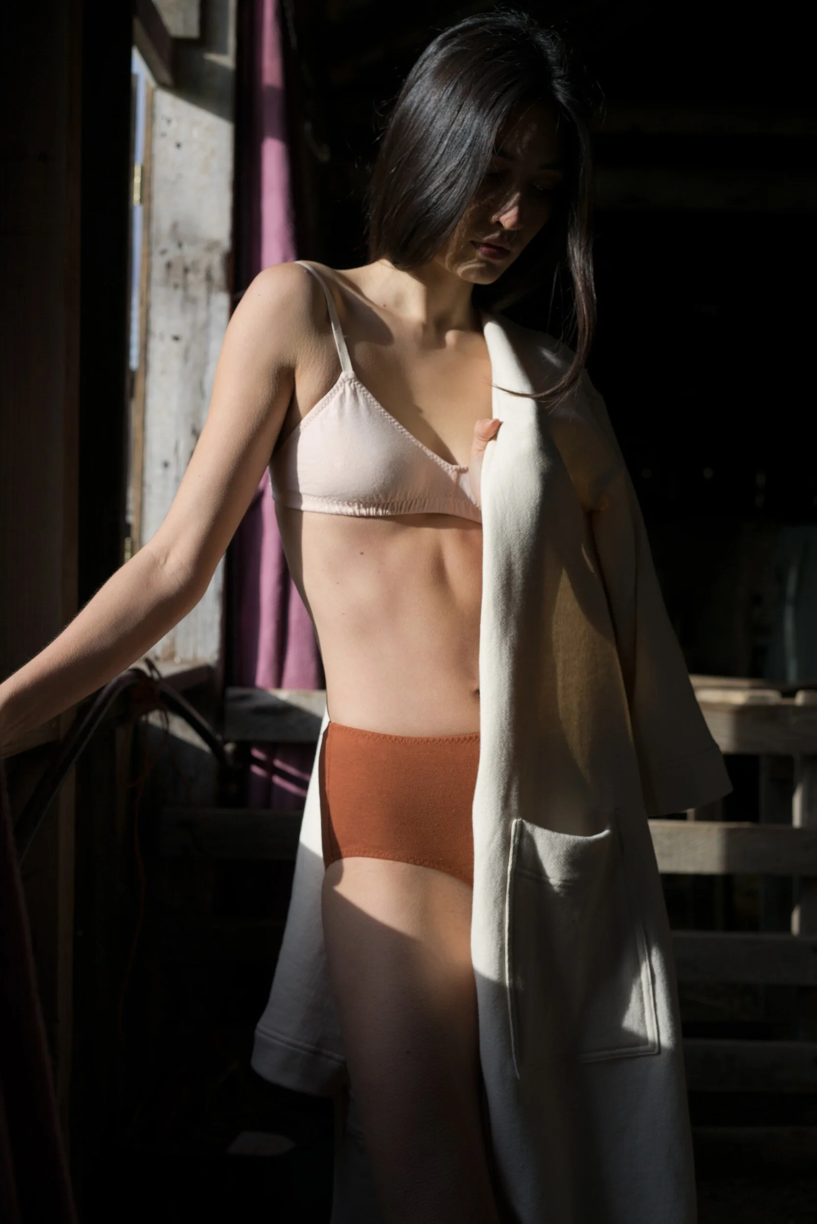 A model in a pale pink bra and rust briefs stands by a barn window with a cream robe draped over one shoulder.