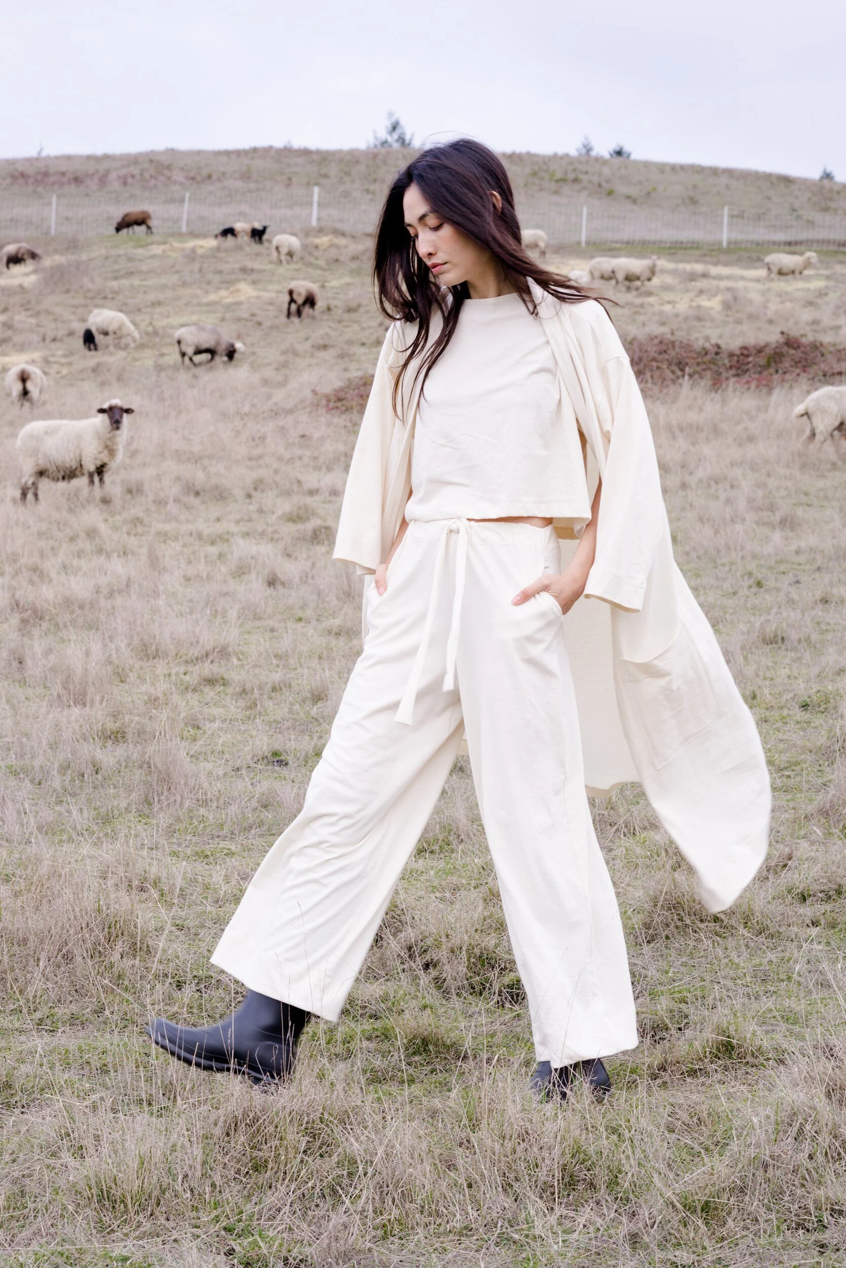 A model in a cream top, trousers and open robe stands with hands in their pockets among sheep in a grassy field.