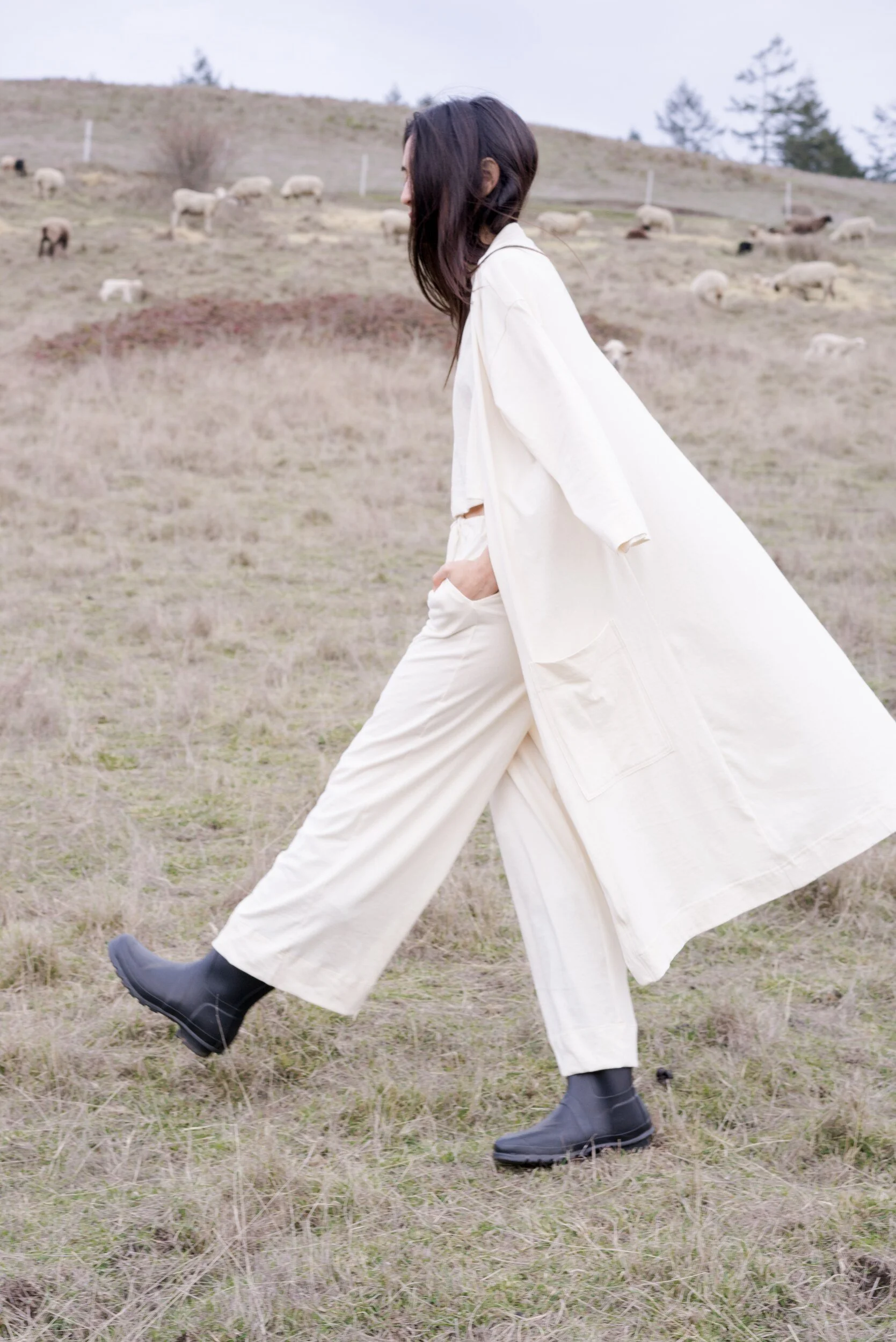 A model in cream clothes and black boots strides across a field, with a long robe trailing behind and sheep grazing nearby.