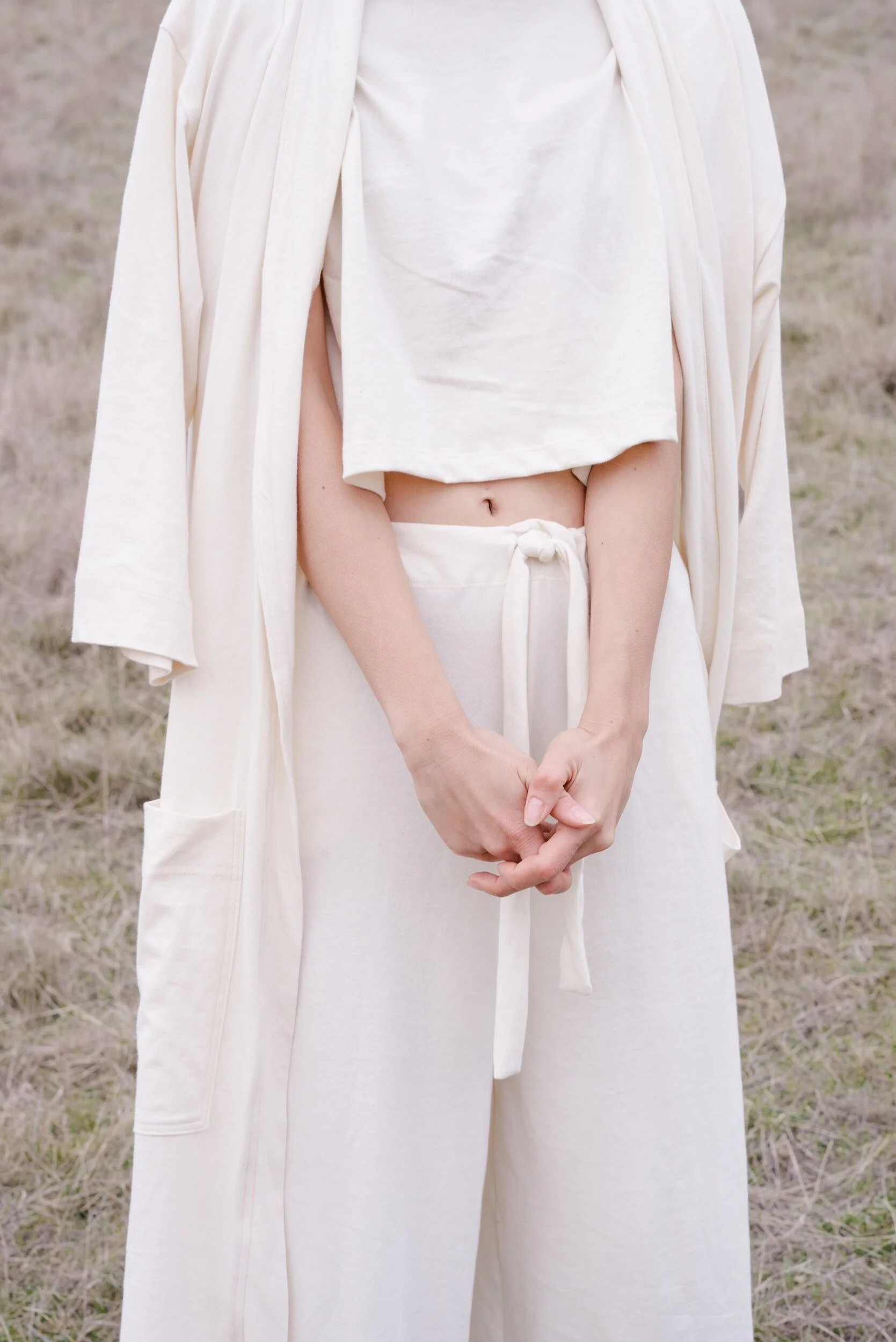 A close view of a model’s clasped hands, cream crop top and drawstring trousers beneath an open robe.