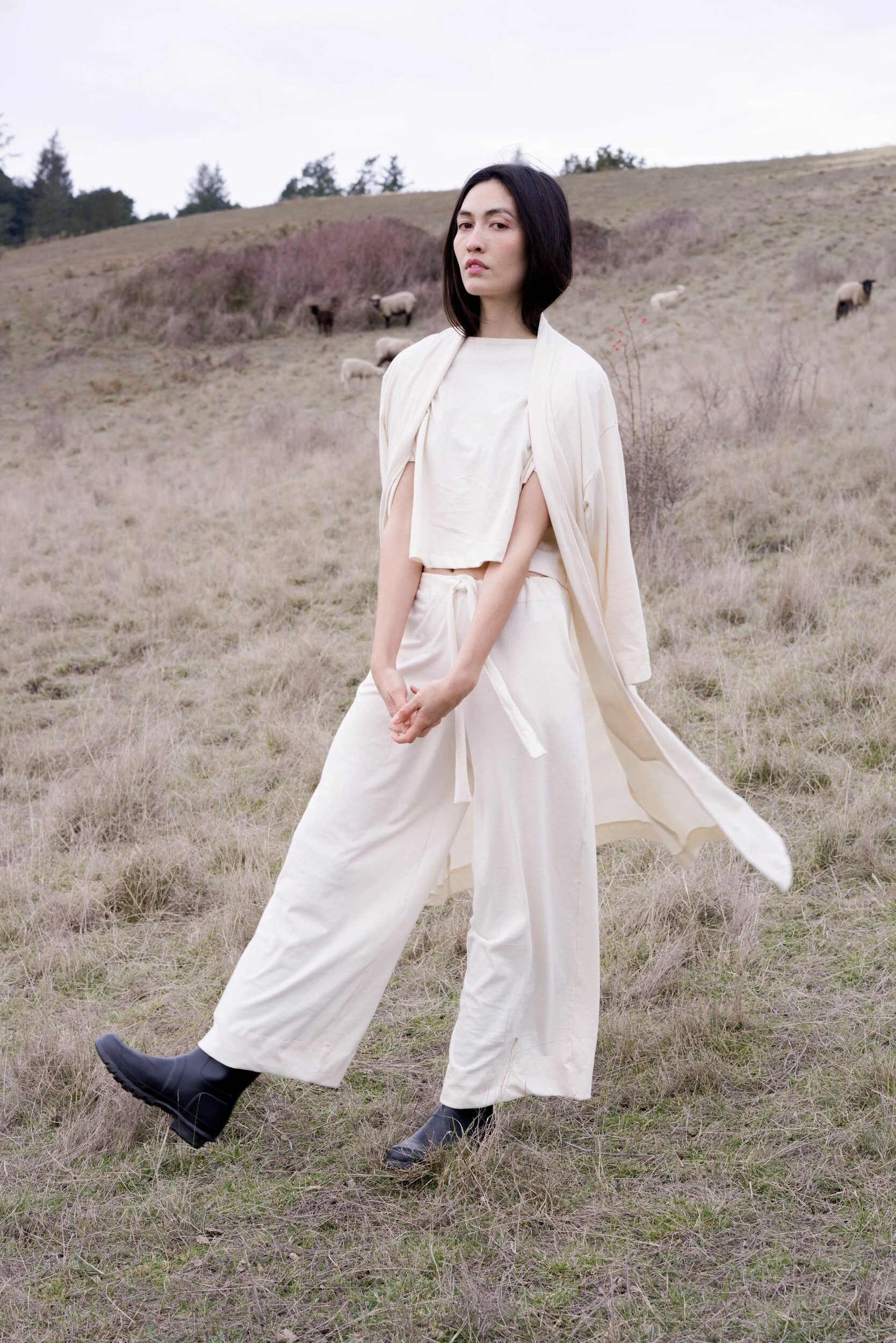 A model in cream clothes and black boots stands with hands clasped and one foot forward on a grassy hillside.