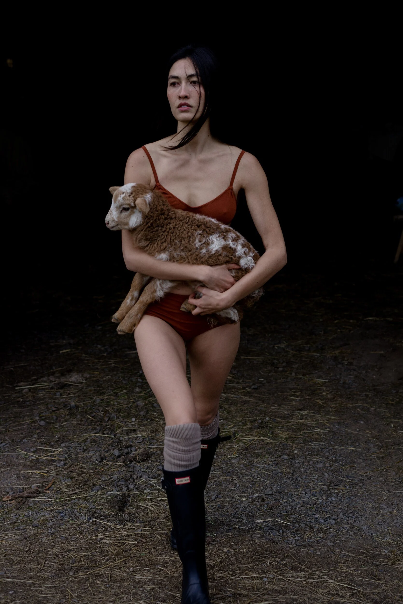A model in rust underwear, thick socks and tall black boots carries a brown and white lamb outside a dark barn.