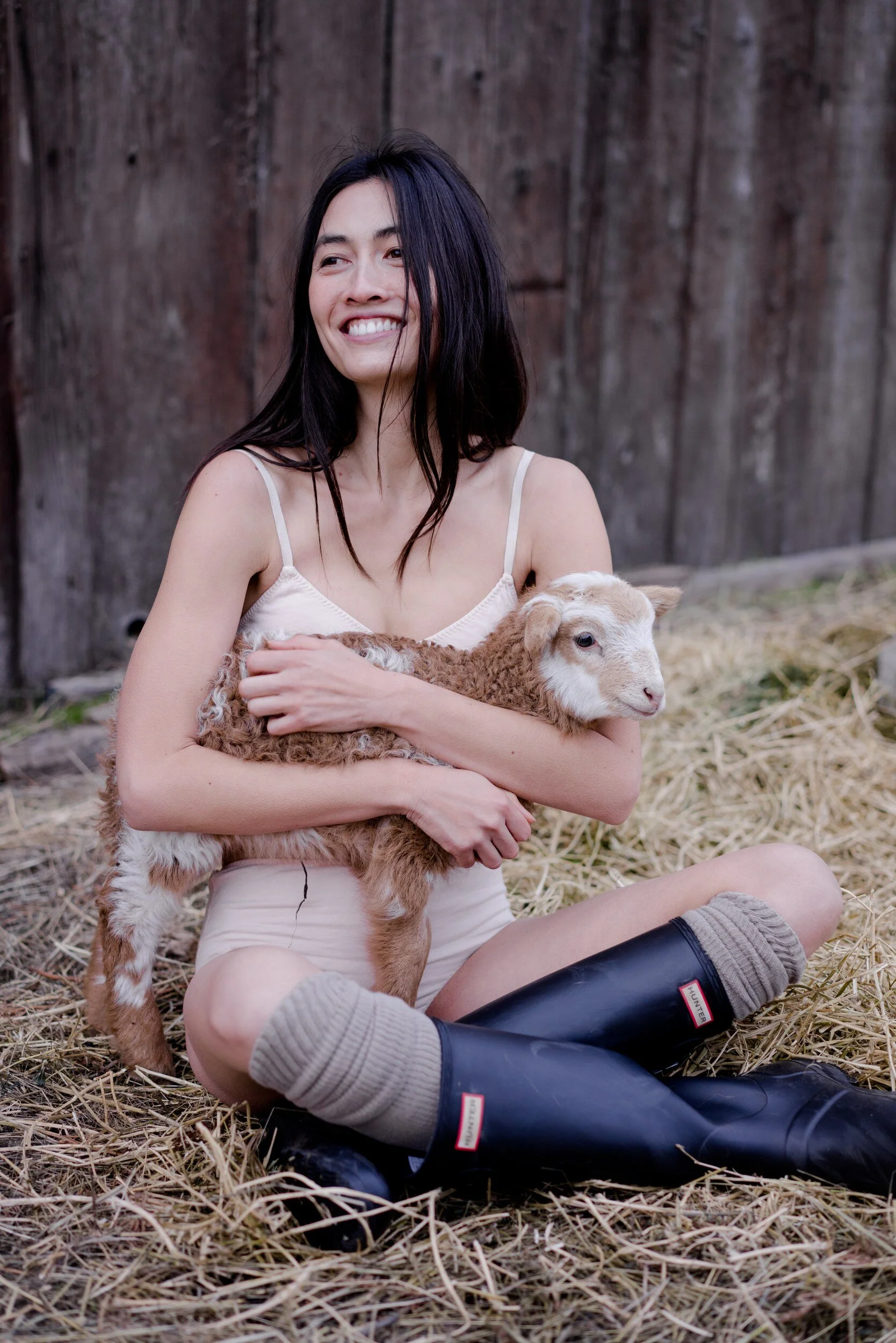 A smiling model in pale pink underwear, thick socks and tall black boots sits on straw with a lamb in their lap.
