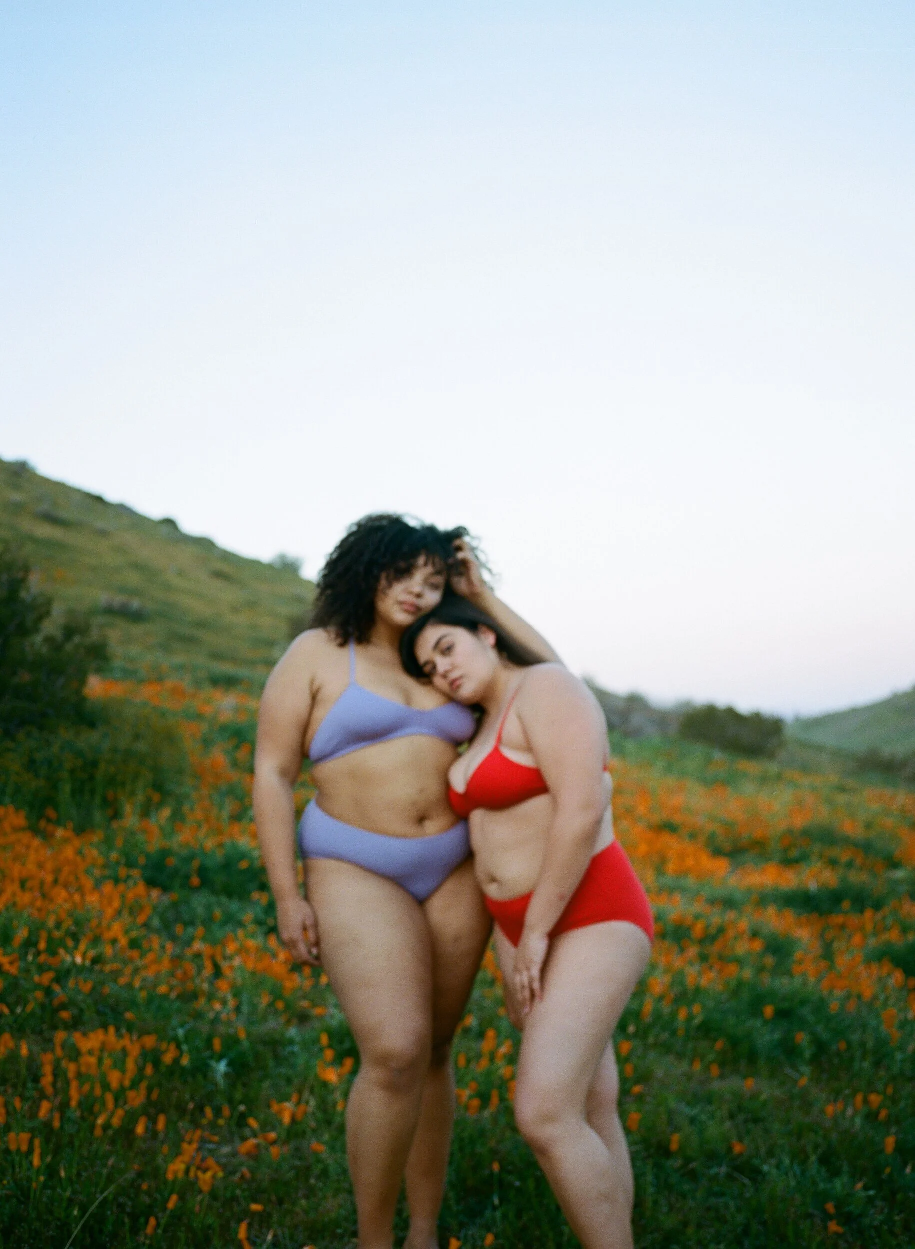 Two models in red and lilac underwear lean together in a field of orange poppies.