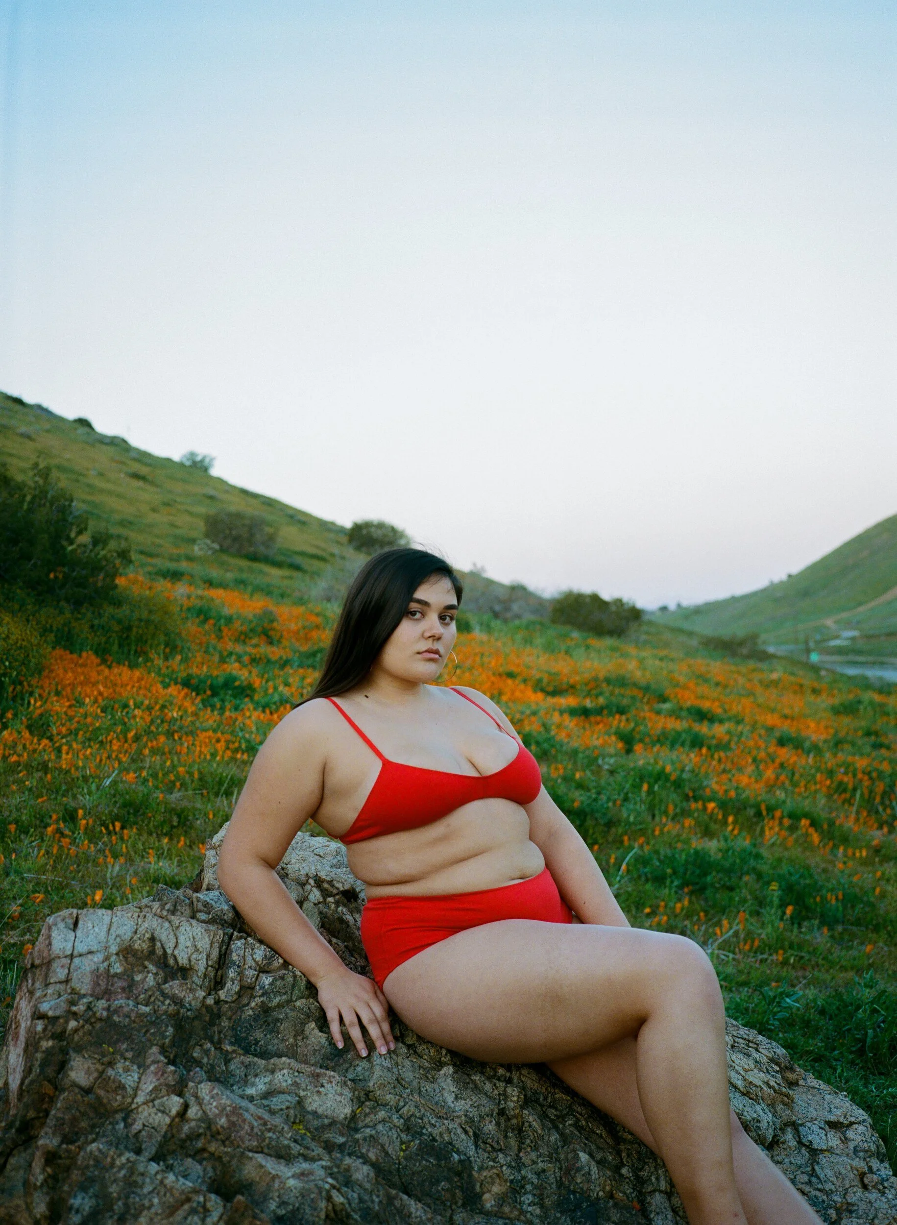 A model in red underwear sits sideways on a rock, with orange poppies covering the hillside behind.