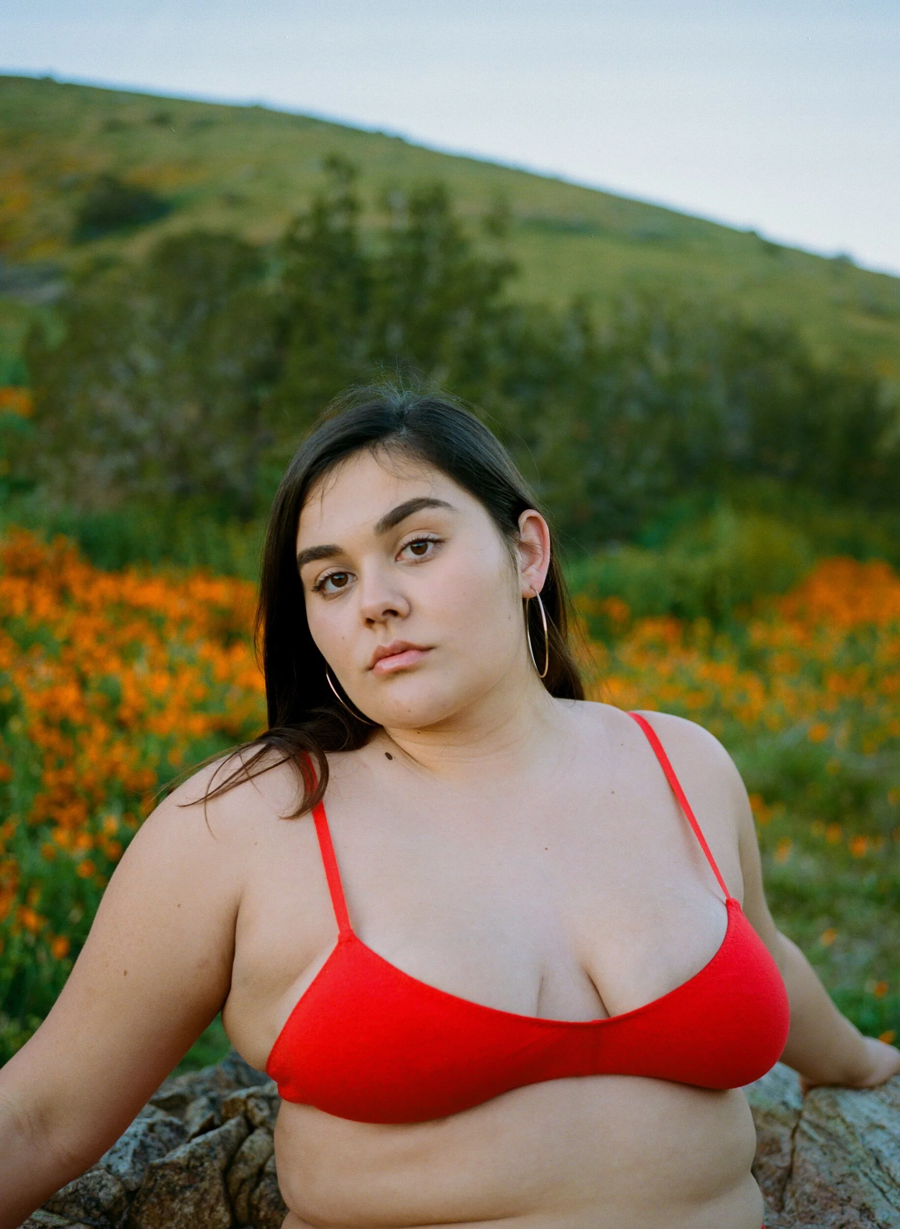 A model in a red bra faces the camera, framed by orange poppies and green hills.