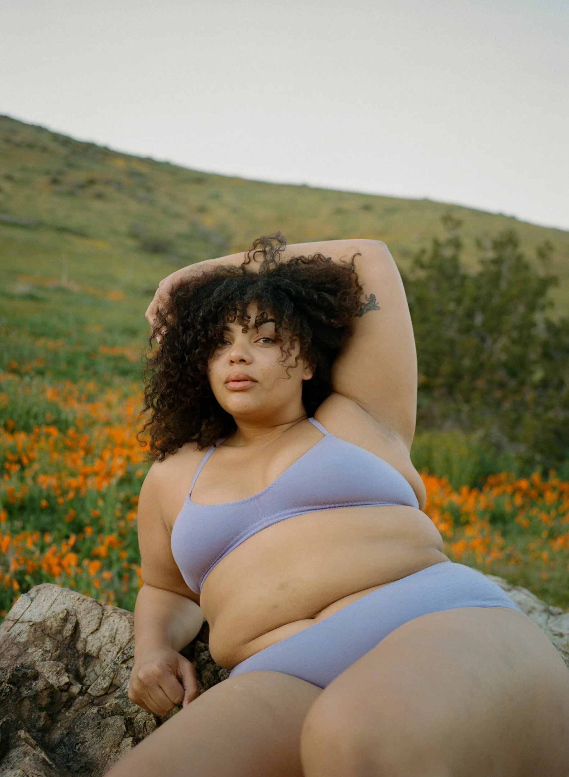 A model in lilac underwear reclines on a rock with one arm raised over their head.