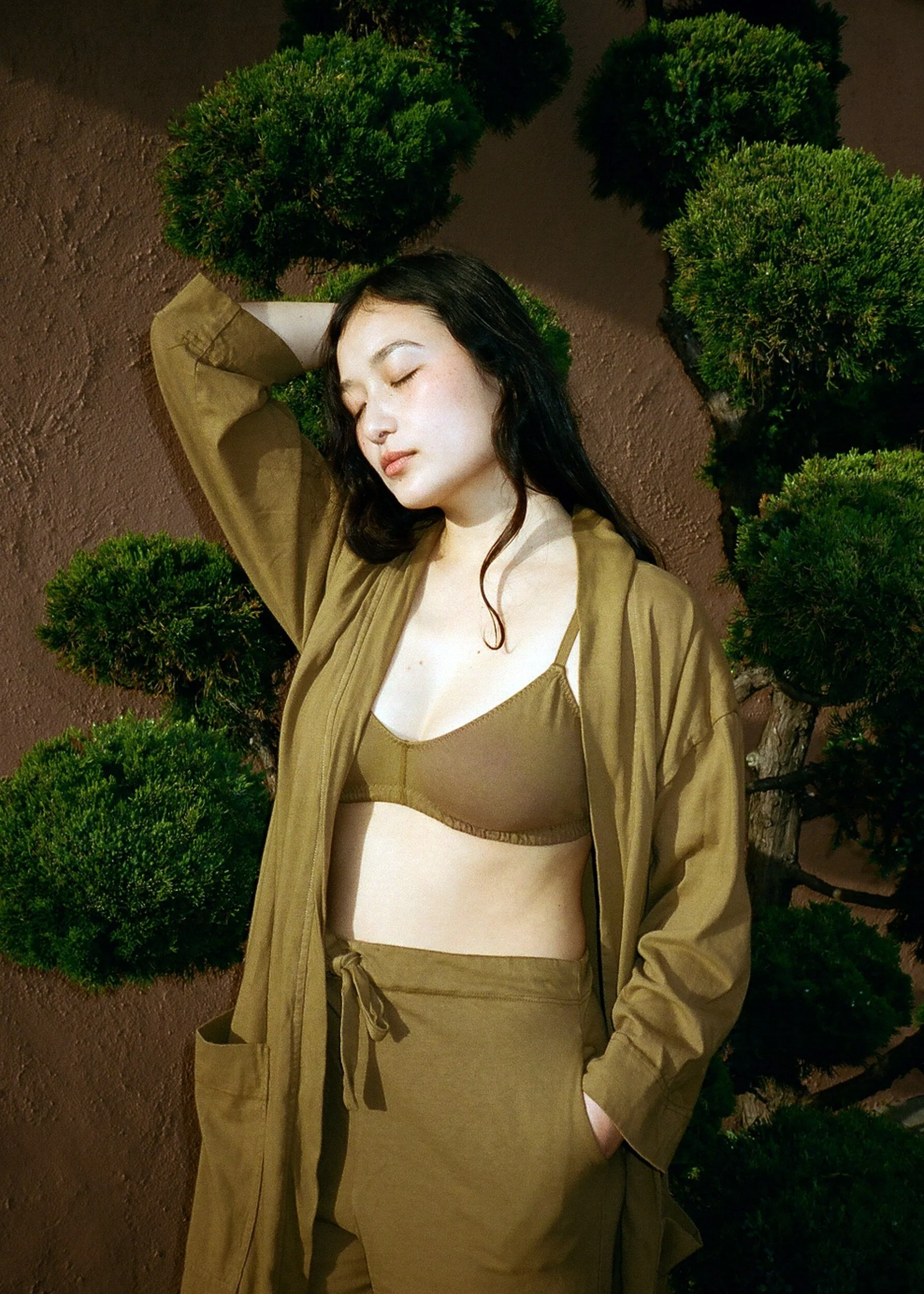 A model in an olive bra, robe and drawstring trousers rests one hand behind their head beside rounded shrubs.