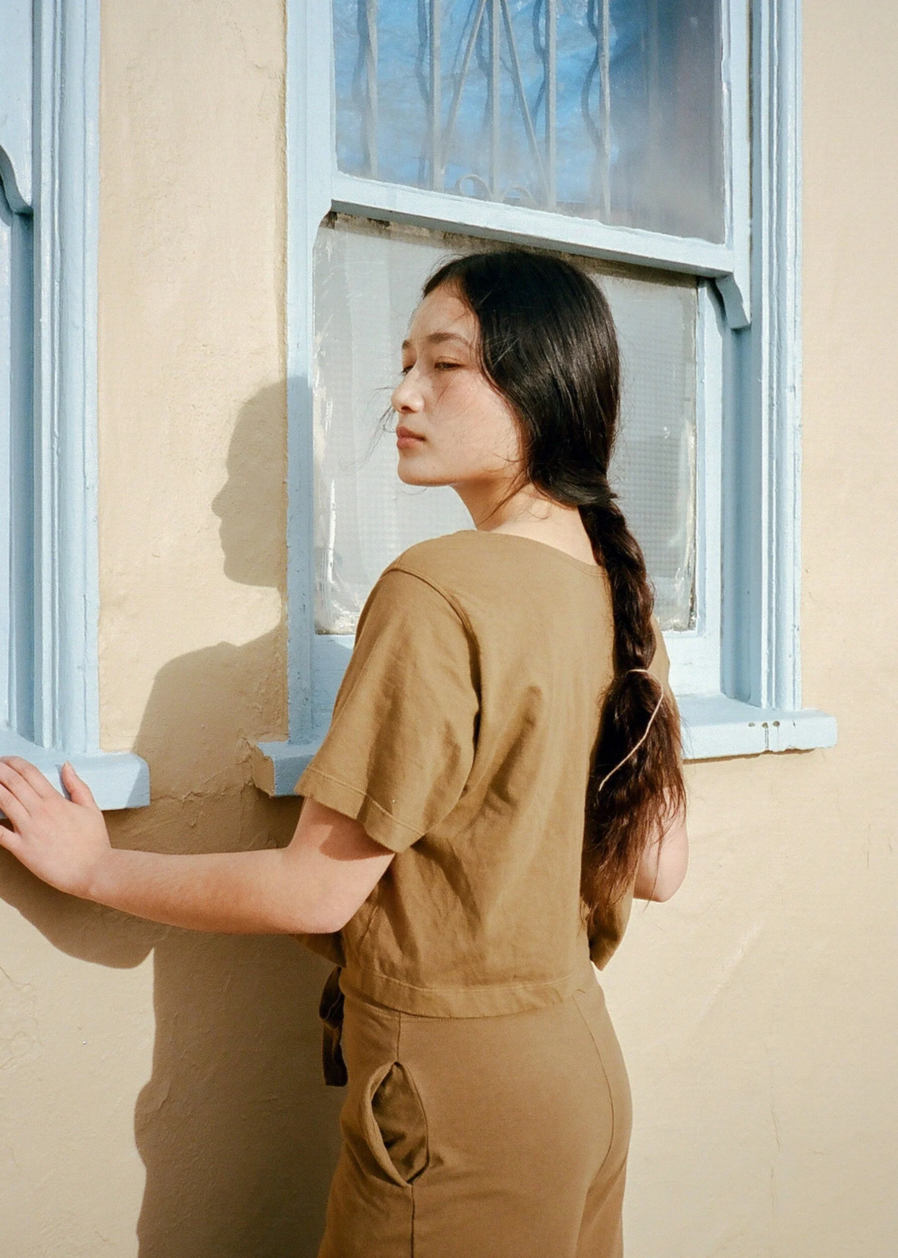 A model in a brown T-shirt and trousers looks over one shoulder beside a pale blue window frame.
