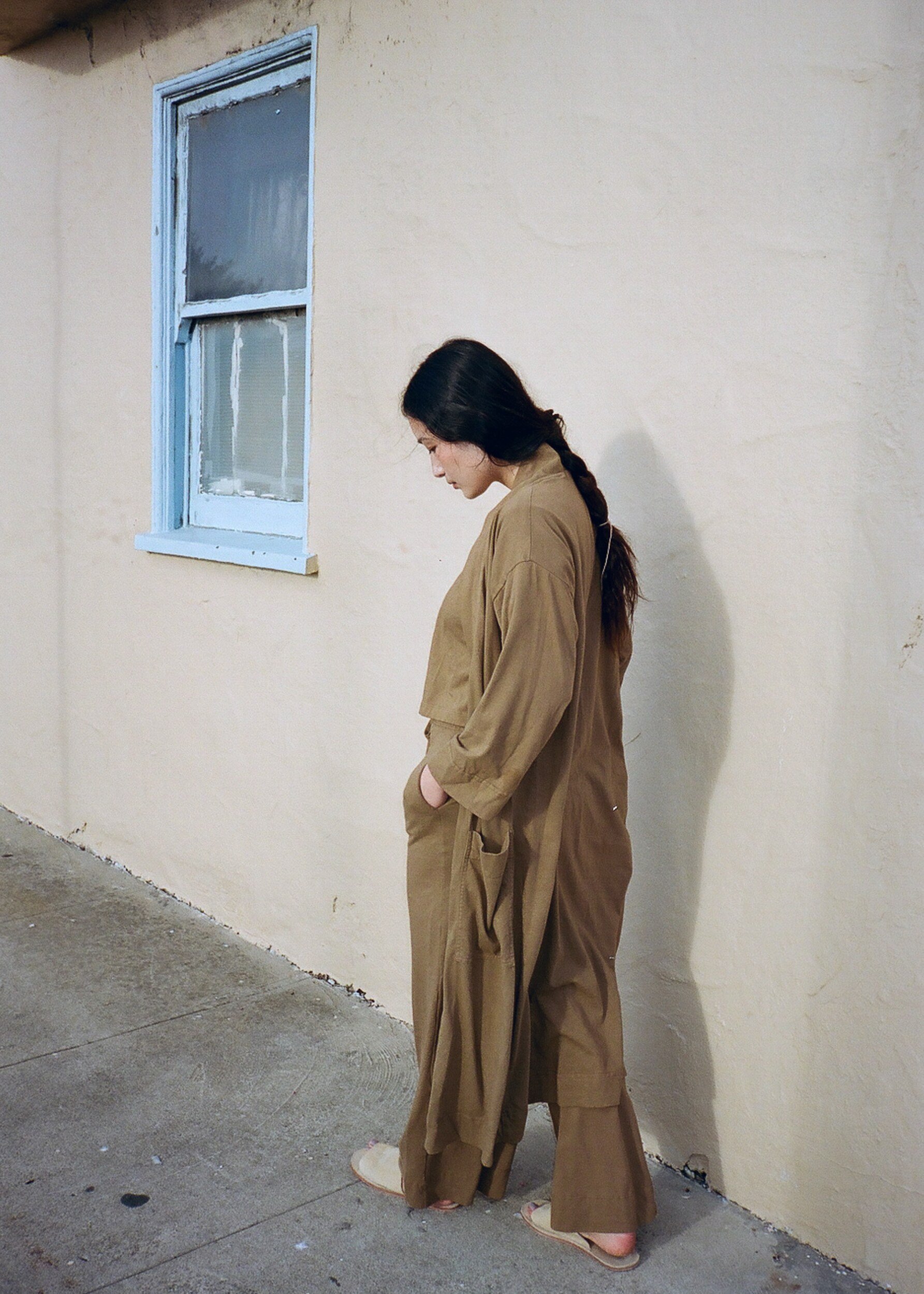 A model in a brown robe and trousers stands beside a pale wall, looking down with one hand in a pocket.
