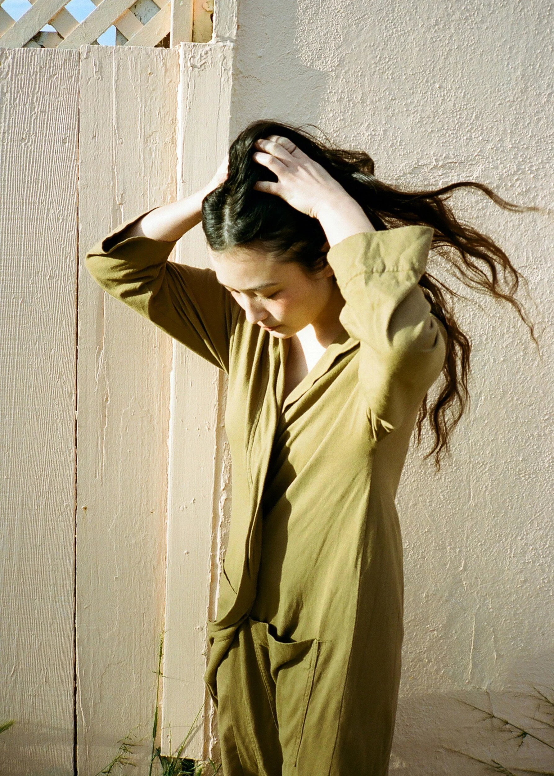 A model in an olive robe lifts their long hair with both hands beside a sunlit pale wall.